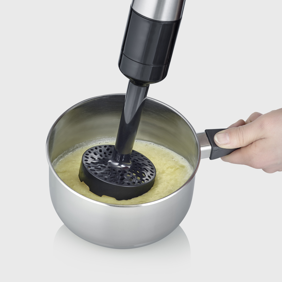 Een staafmixer in een pan, die een gele mix mixt. De pan is van roestvrij staal.