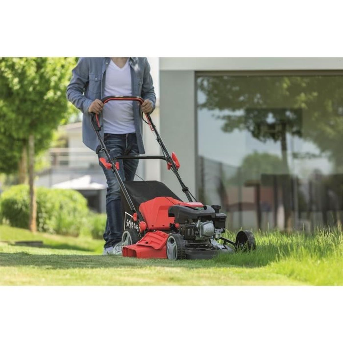 Een persoon maait een gazon met een rood-zwarte grasmaaier, met bomen en een huis op de achtergrond.