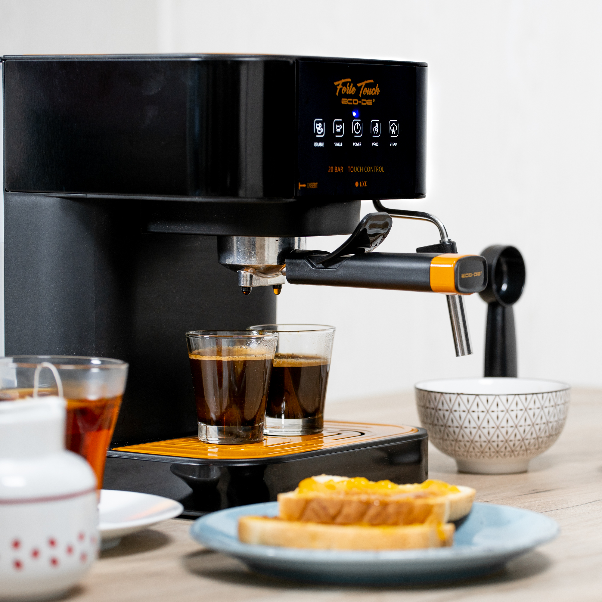 Máquina de espresso con dos vasos de chupito, tostadas y un cuenco sobre una superficie de madera.