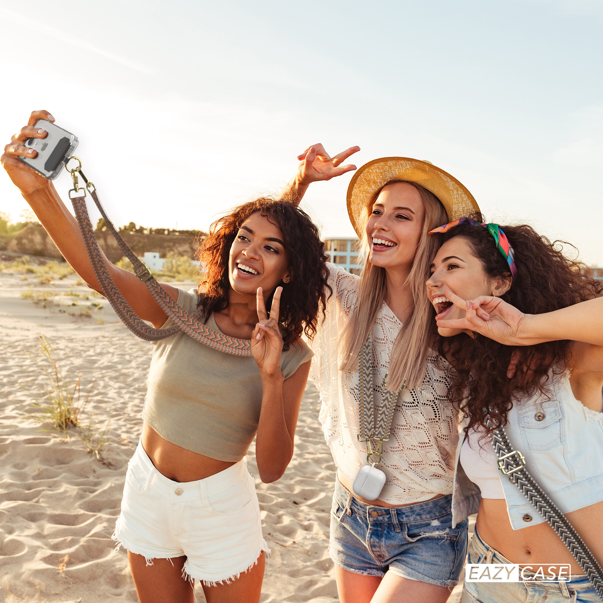 Drei Frauen machen ein Selfie am Strand, benutzen ein Telefon mit Gurt. Fröhliche Ausdrücke und sonnige Atmosphäre.
