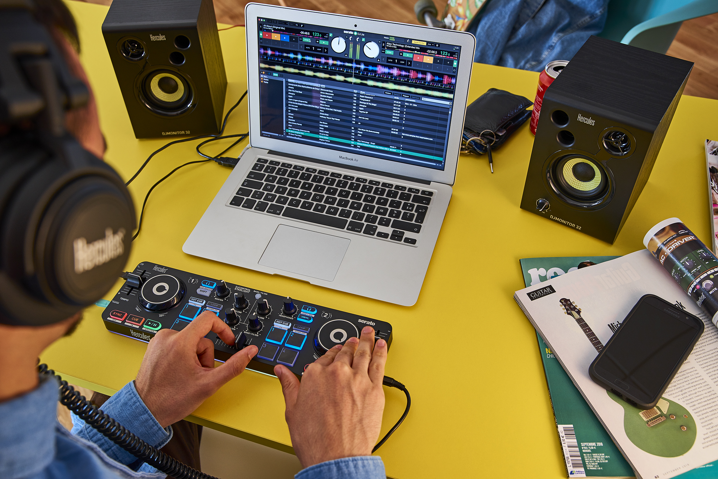 Een persoon die DJ-apparatuur gebruikt. Er staan luidsprekers, een laptop en een mengpaneel op een gele tafel.