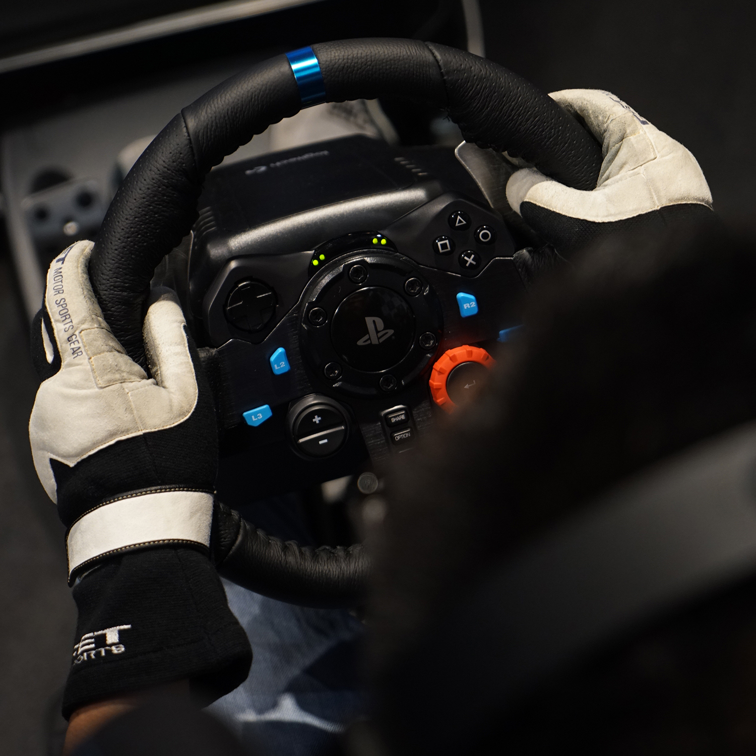 Hände mit Handschuhen halten ein schwarzes Lenkrad mit Knöpfen und PlayStation-Logo.