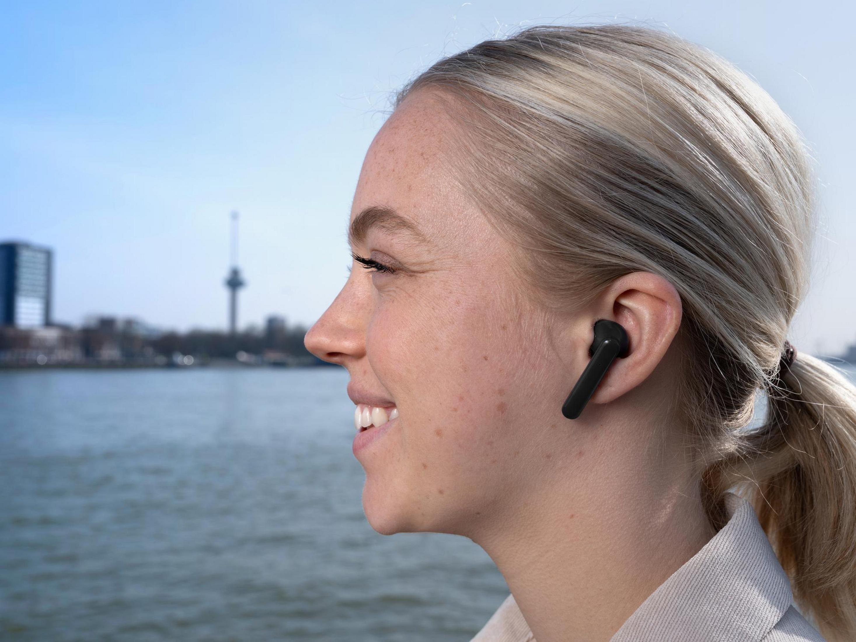 Kobieta z blond włosami nosi czarną słuchawkę, uśmiechając się w pobliżu wody i budynków.