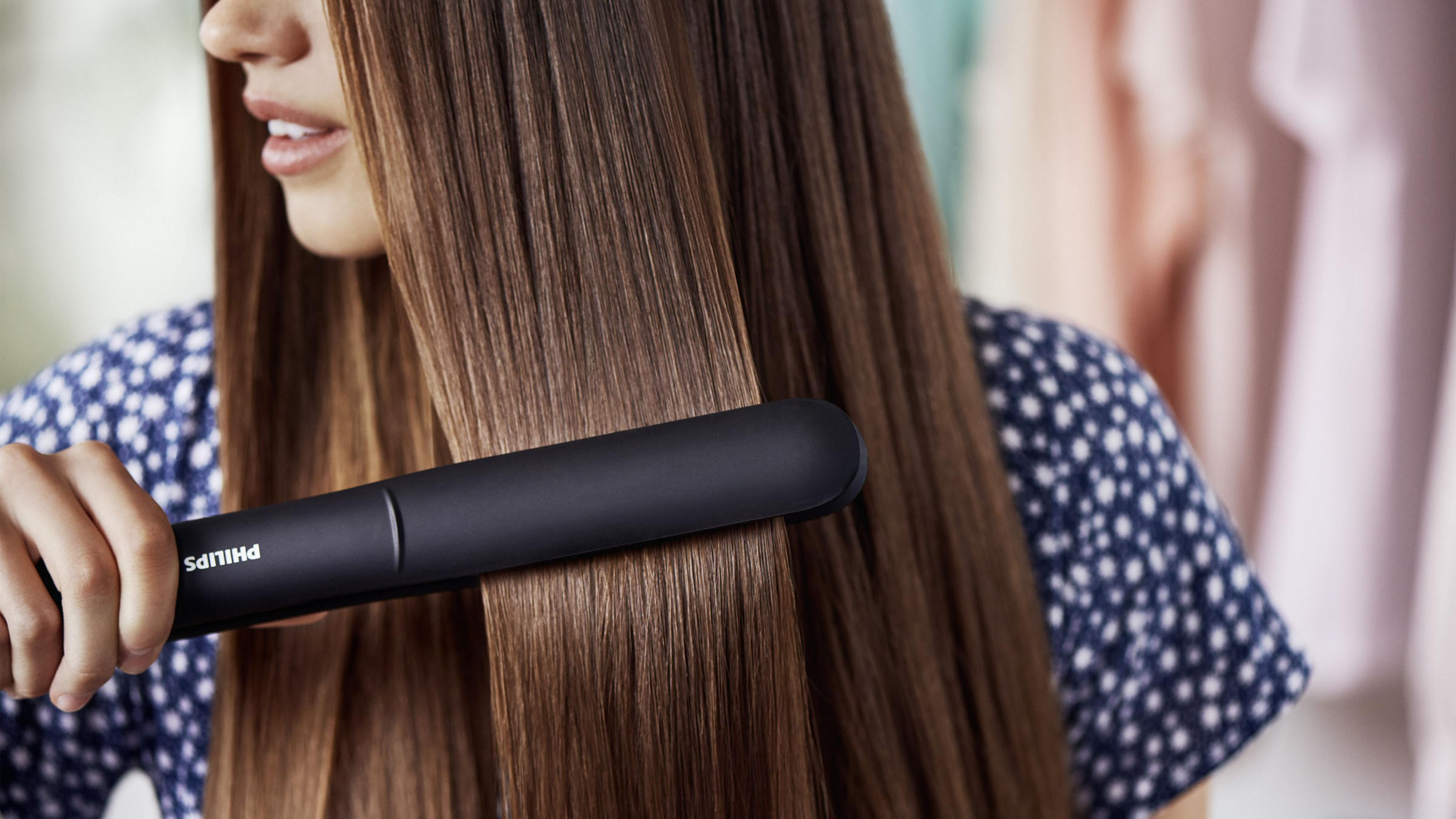 Kobieta używająca czarnej prostownicy do długich, brązowych włosów. Ma na sobie koszulę w kropki.