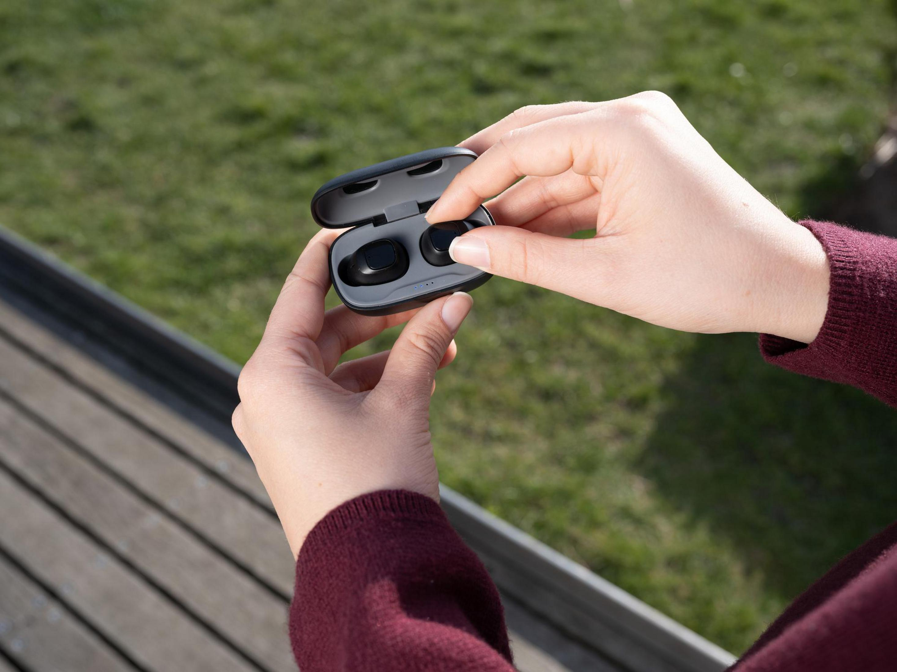 Dłonie trzymające czarno-szare etui na słuchawki, wyjmujące czarną słuchawkę.