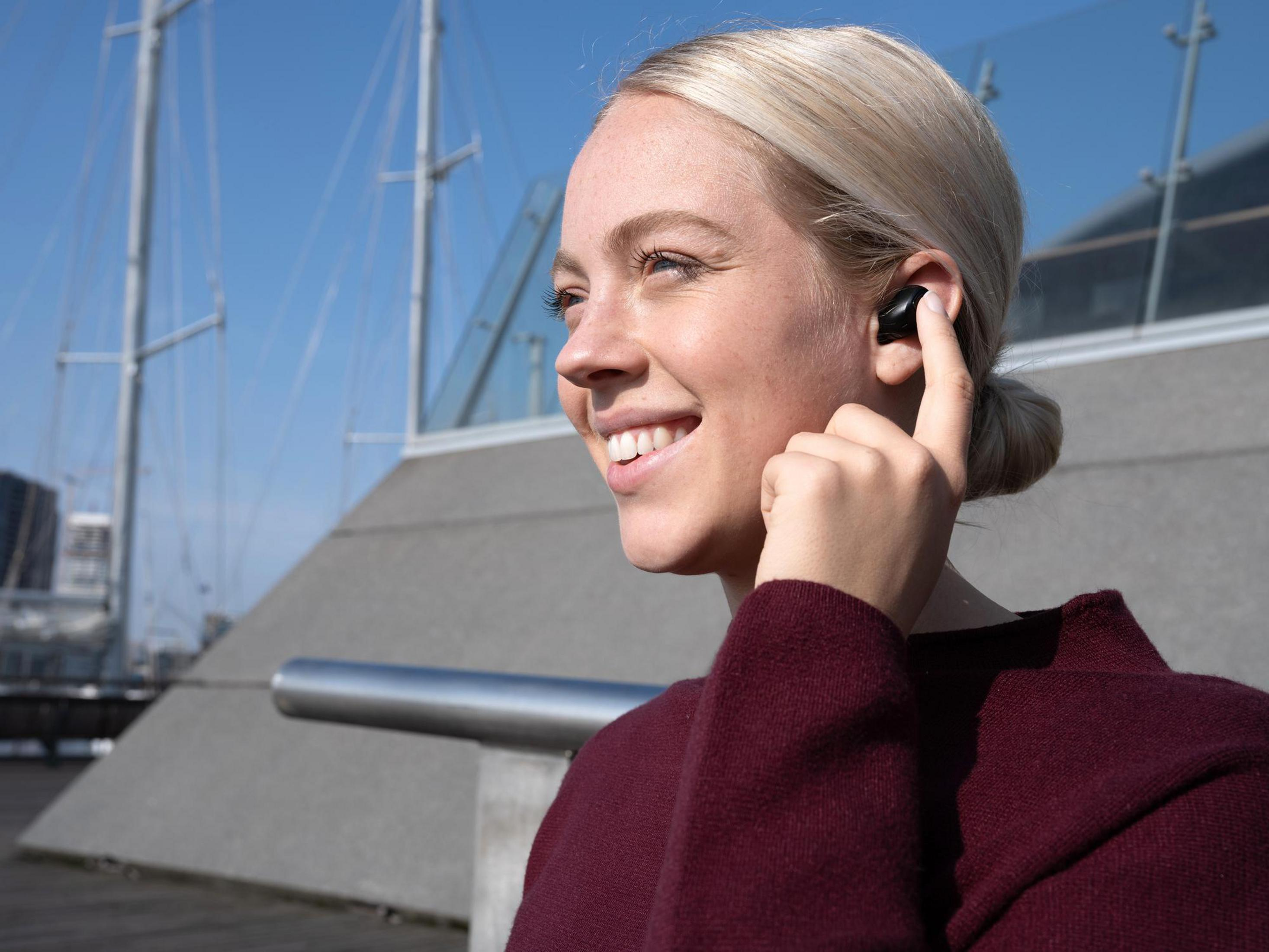 Kobieta z blond włosami nosząca słuchawkę i uśmiechająca się. Na zewnątrz z błękitnym niebem i budynkiem.