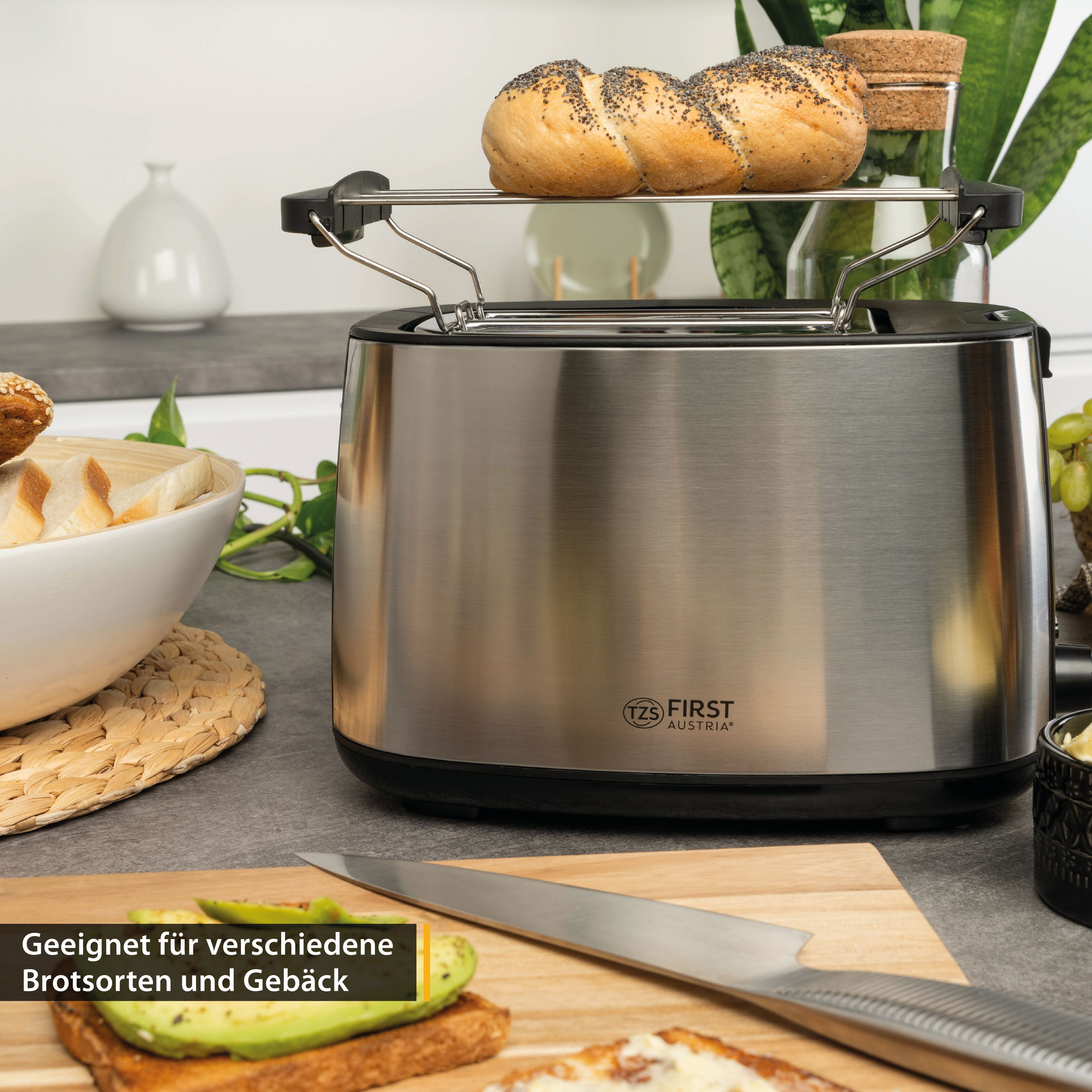 Ein Toaster aus Edelstahl mit zwei Brötchen auf dem oberen Rost und Brot und Avocado auf dem Schneidebrett.