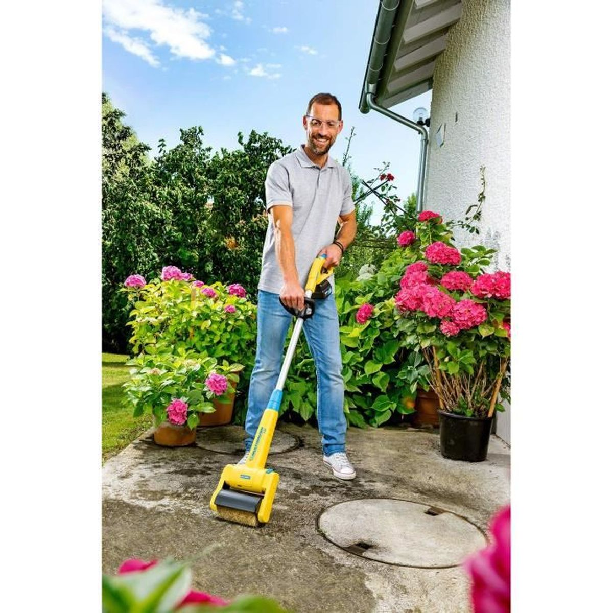 Ein Mann benutzt ein gelb-blaues Walzwerkzeug auf einer Betonoberfläche. Topfpflanzen mit rosa Blüten sind im Hintergrund.
