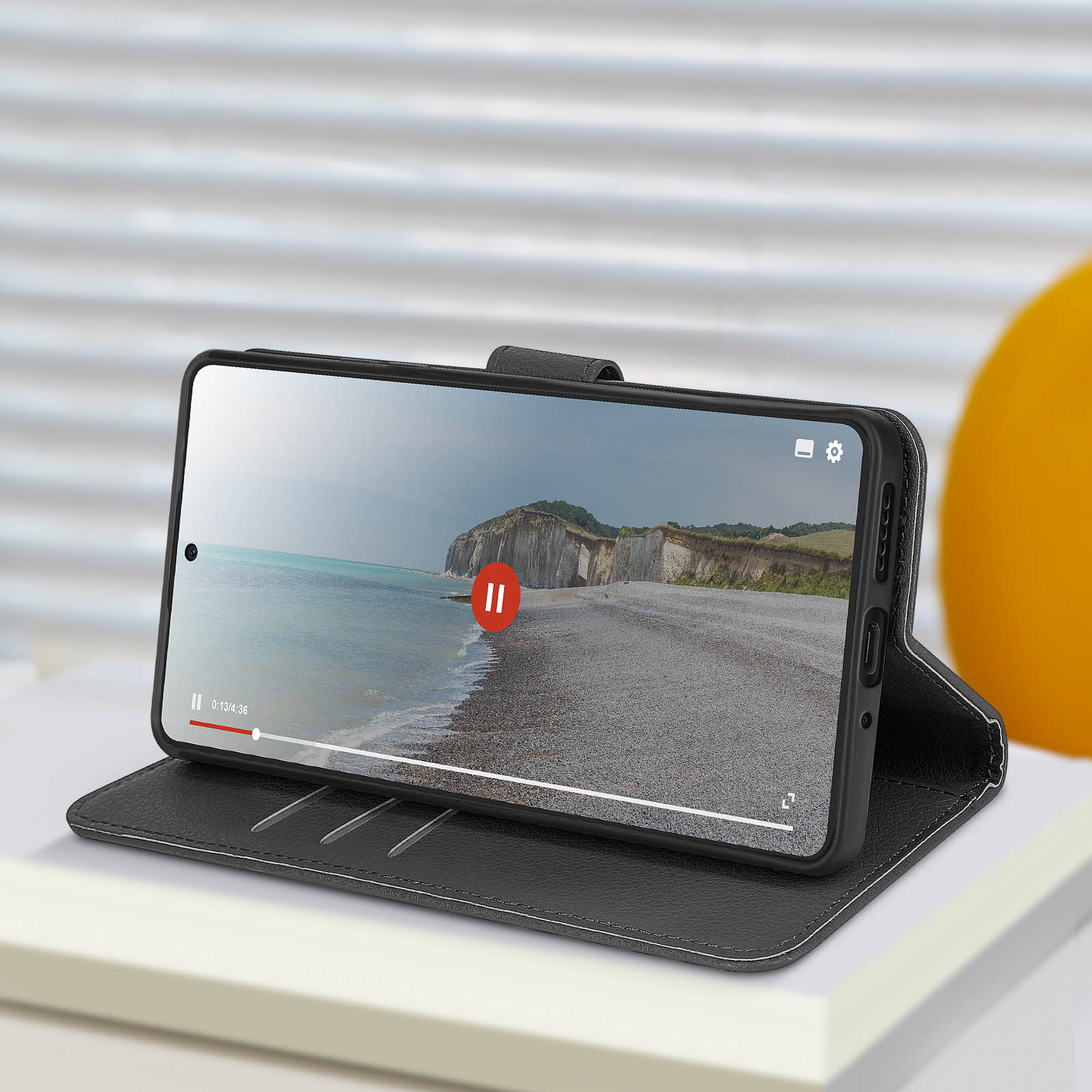 Un telefono che mostra un video di una spiaggia su un libro, in una custodia nera. Un tavolo bianco e un oggetto giallo sullo sfondo.