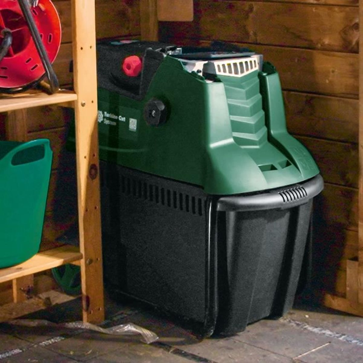 Une poubelle à compost verte à l'intérieur d'un hangar en bois. La poubelle est noire en bas et a une poignée rouge en haut.