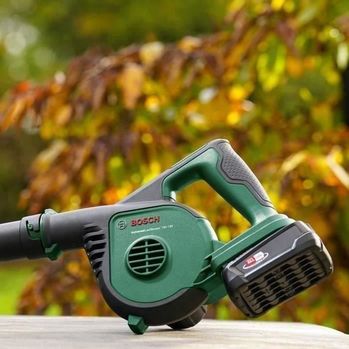 Zielona i czarna dmuchawa do liści, trzymana na tle rozmytych liści.