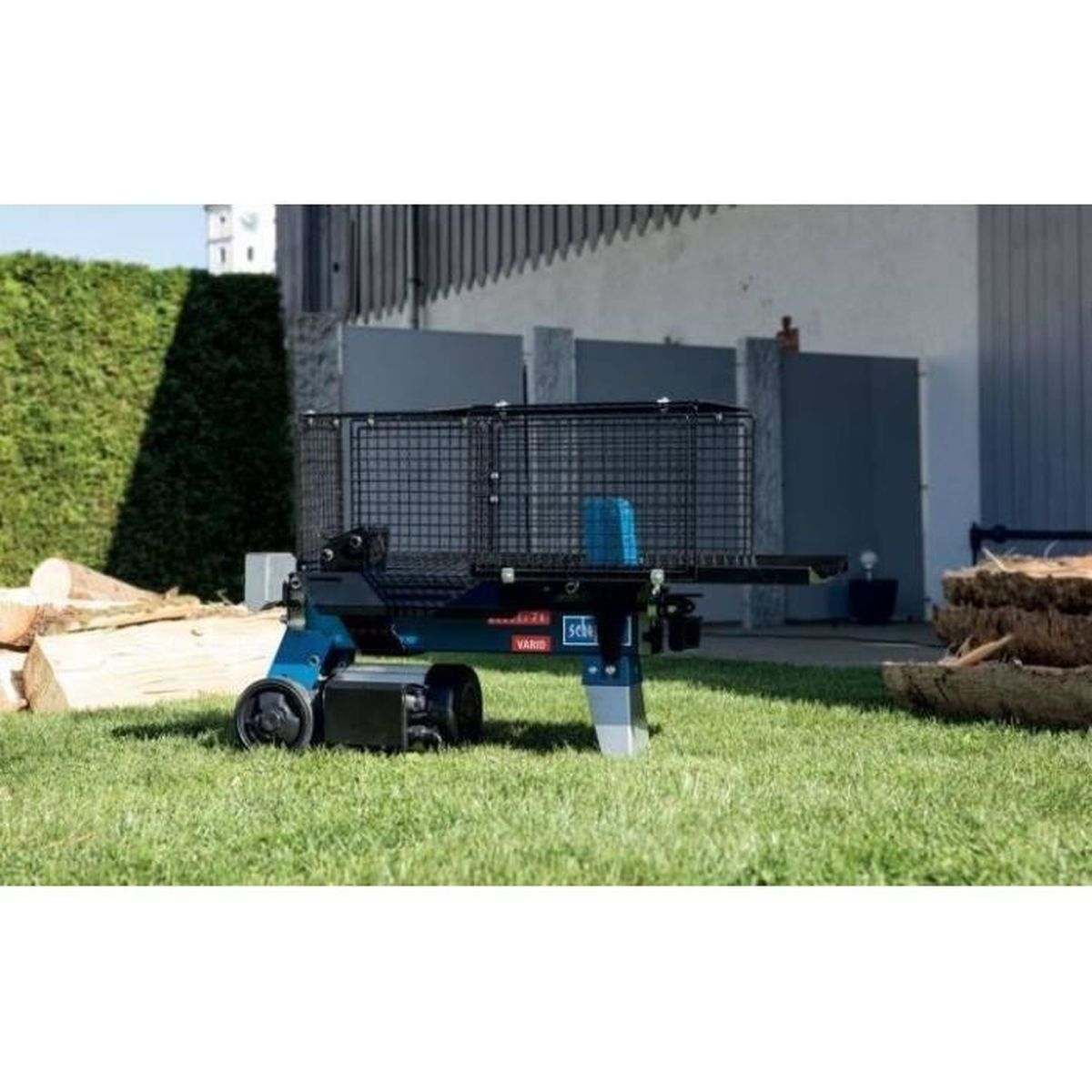 Eine blau-schwarze Holzspaltmaschine auf Gras mit Holz und Gebäude im Hintergrund.