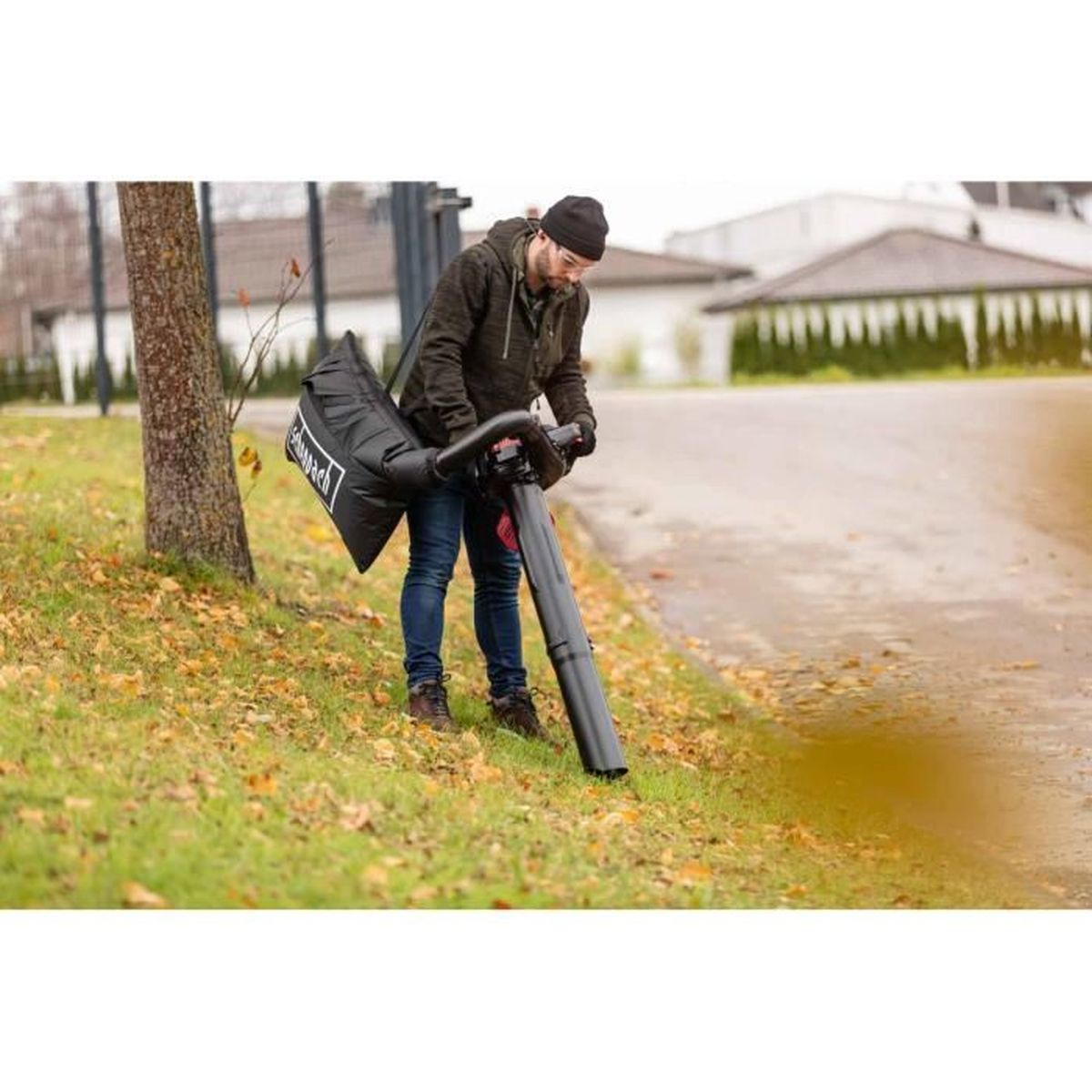 Mężczyzna w czarnym kapeluszu i płaszczu używa dmuchawy do liści na trawniku z liśćmi.