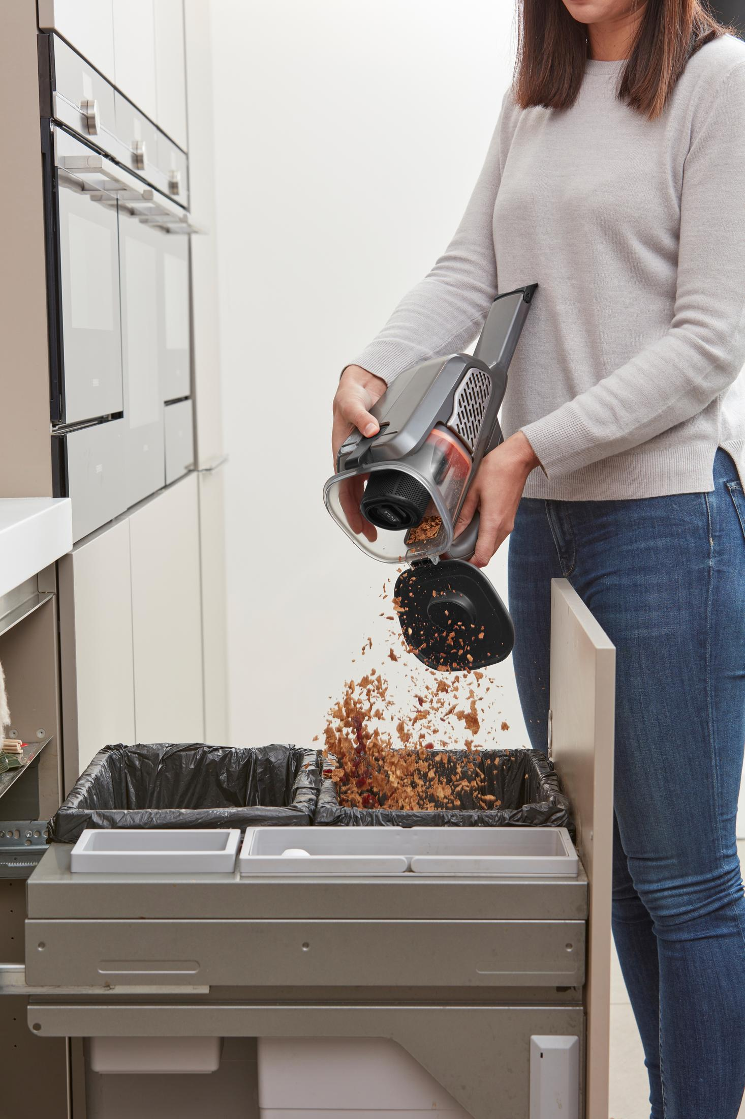 Woman emptying a handheld vacuum into a double trash bin in a kitchen setting.