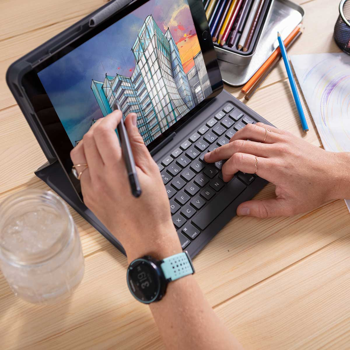 Persoon typt op een tablet toetsenbord, tekent op het scherm, een horloge om de pols en potloden op tafel.
