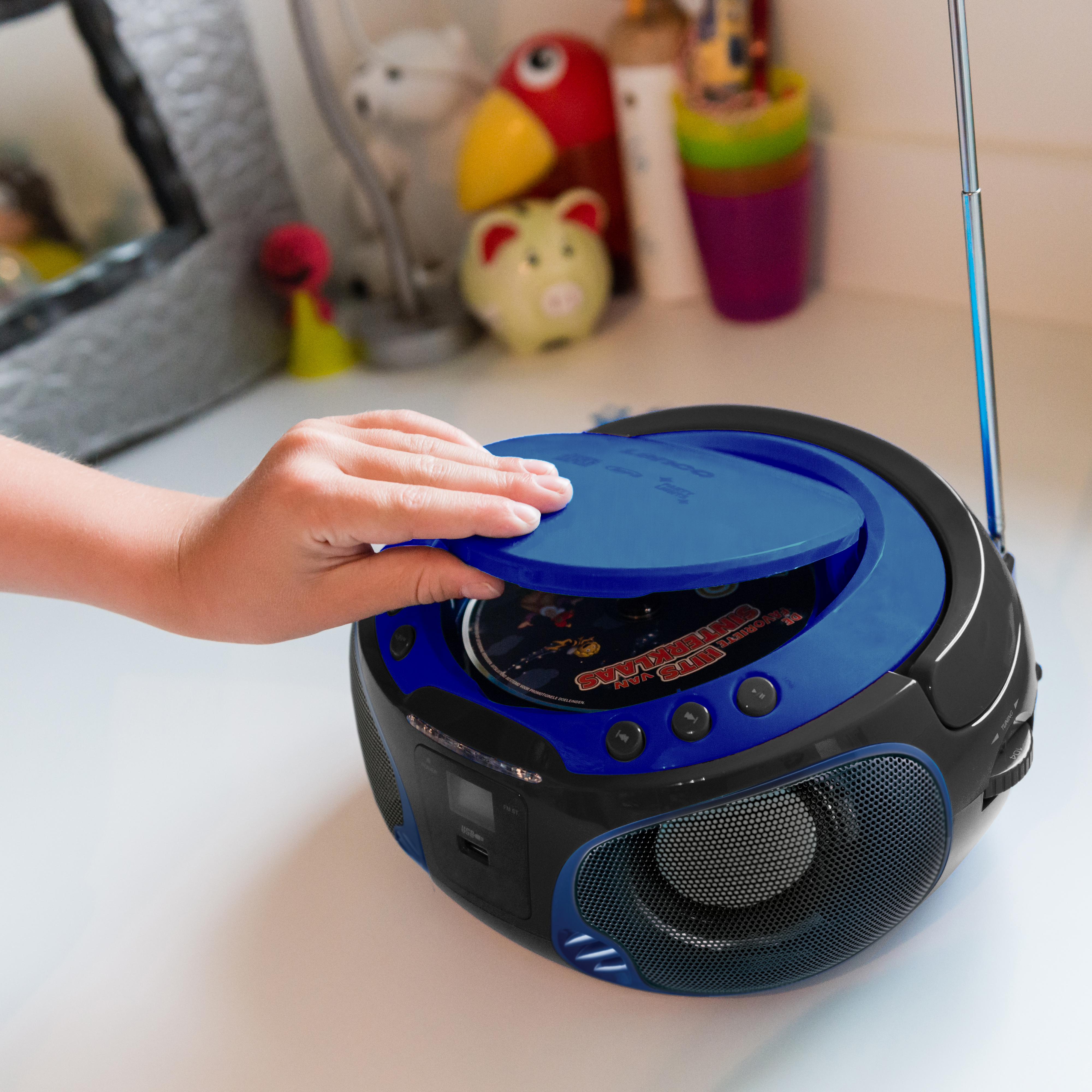 Child's hand opening a blue CD player on white surface, toys in background.