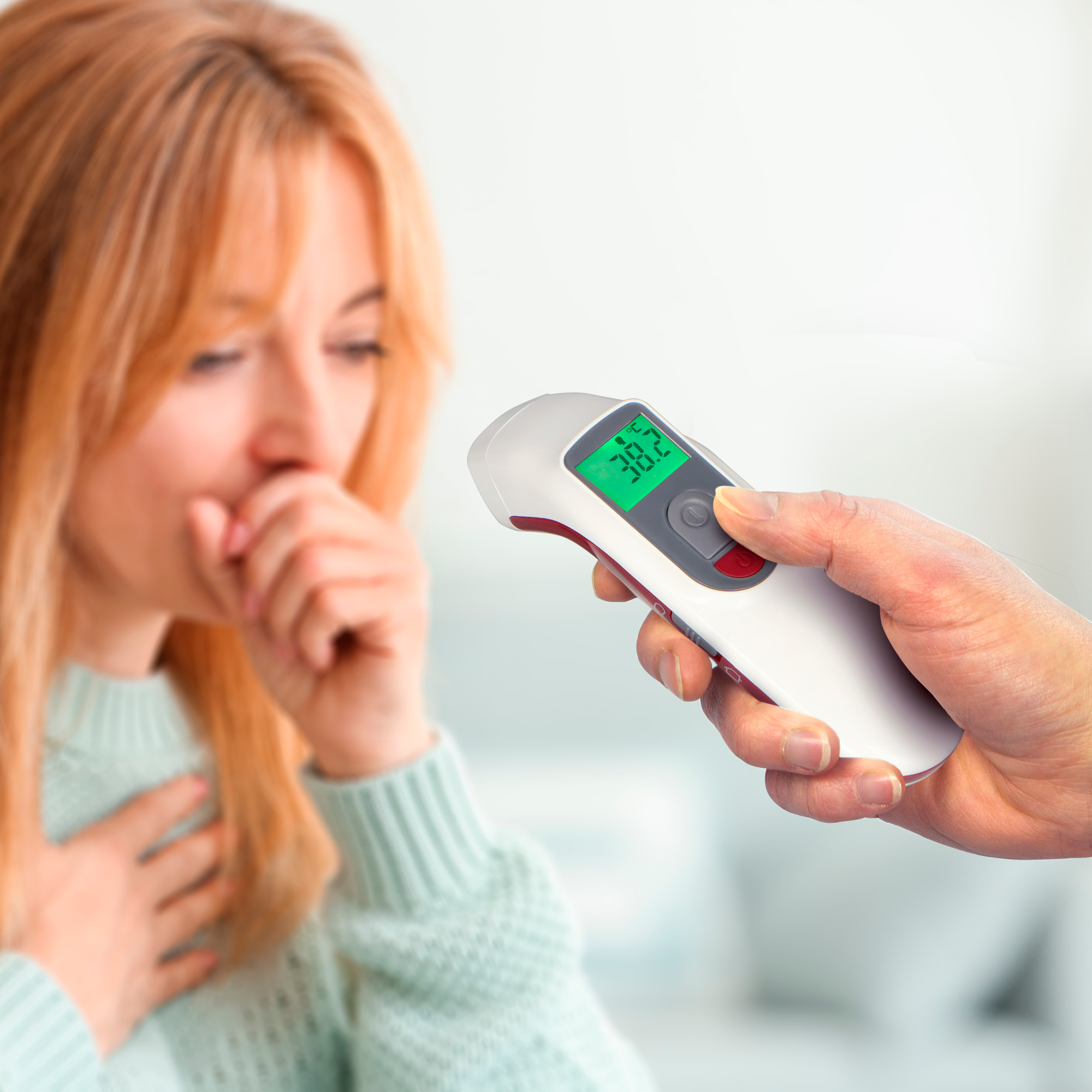 Die Hand einer Person hält ein Thermometer an eine hustende Frau.