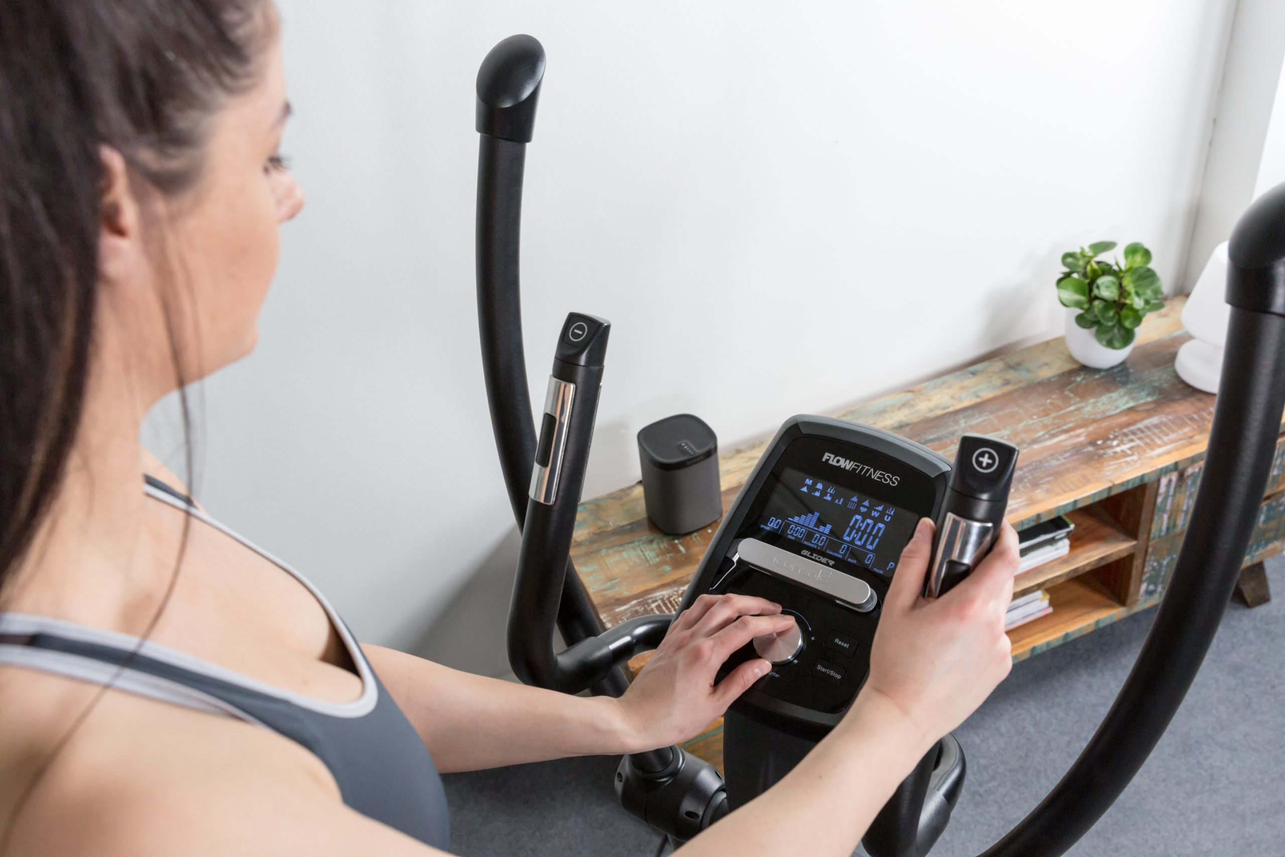 Frau auf einem Crosstrainer. Sie benutzt die Maschine. Hintergrund: Schrank, Pflanze.
