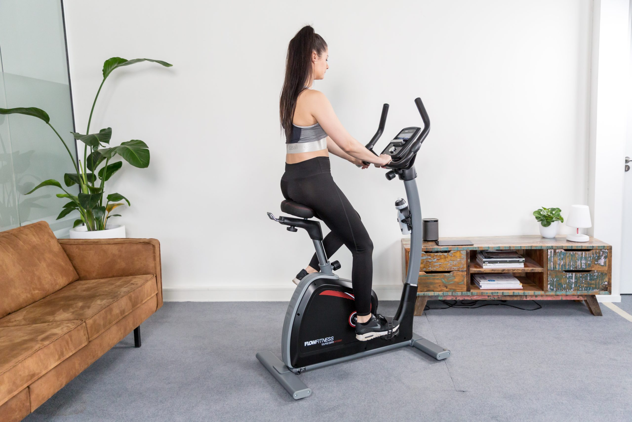 Frau auf einem Heimtrainer. Sie ist in einem Raum mit Sofa, Schrank und Fenster.