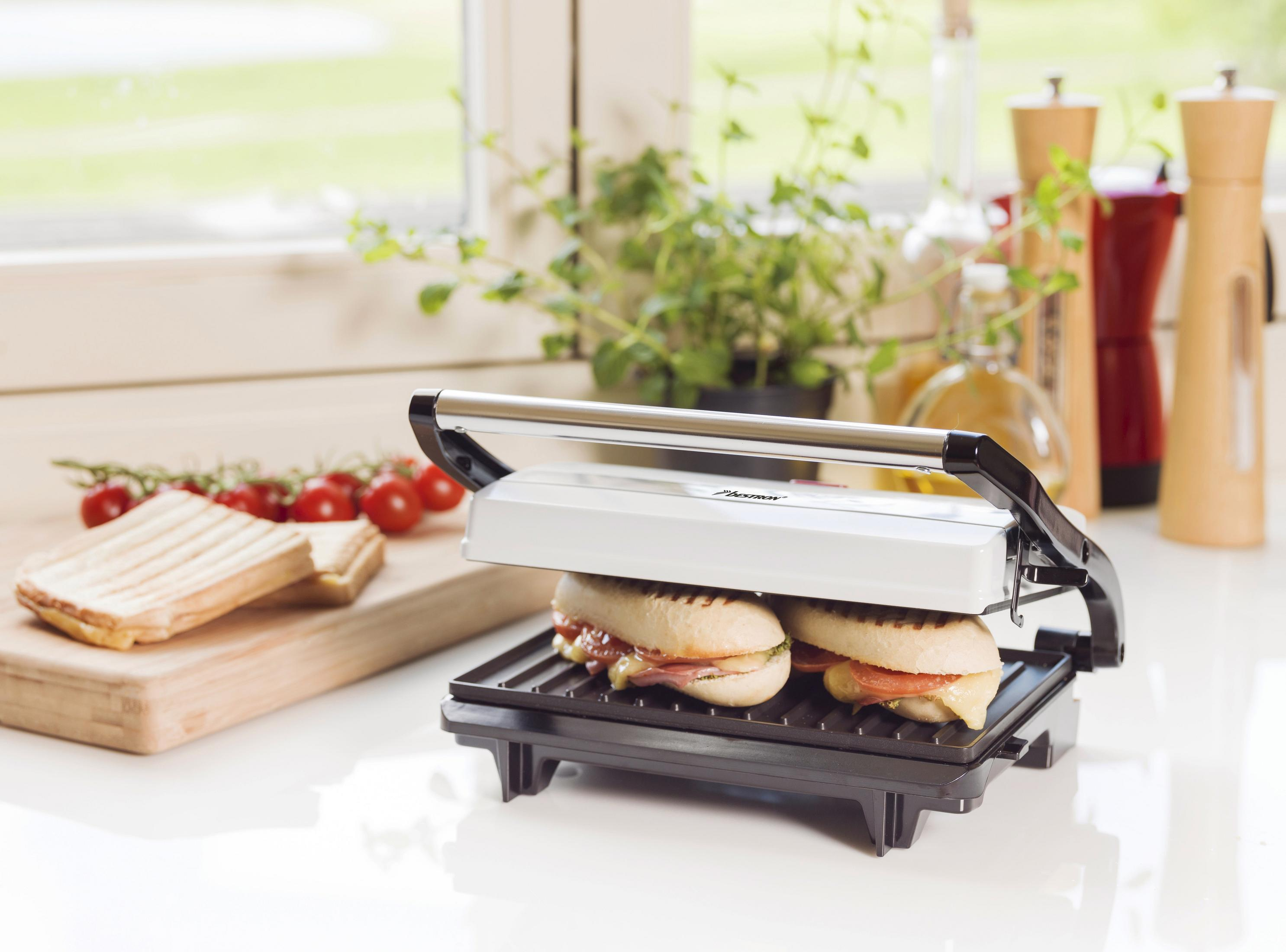 Eine Panini-Presse mit zwei Sandwiches. Die Sandwiches stehen auf einer weißen Theke, mit Tomaten und Kräutern.