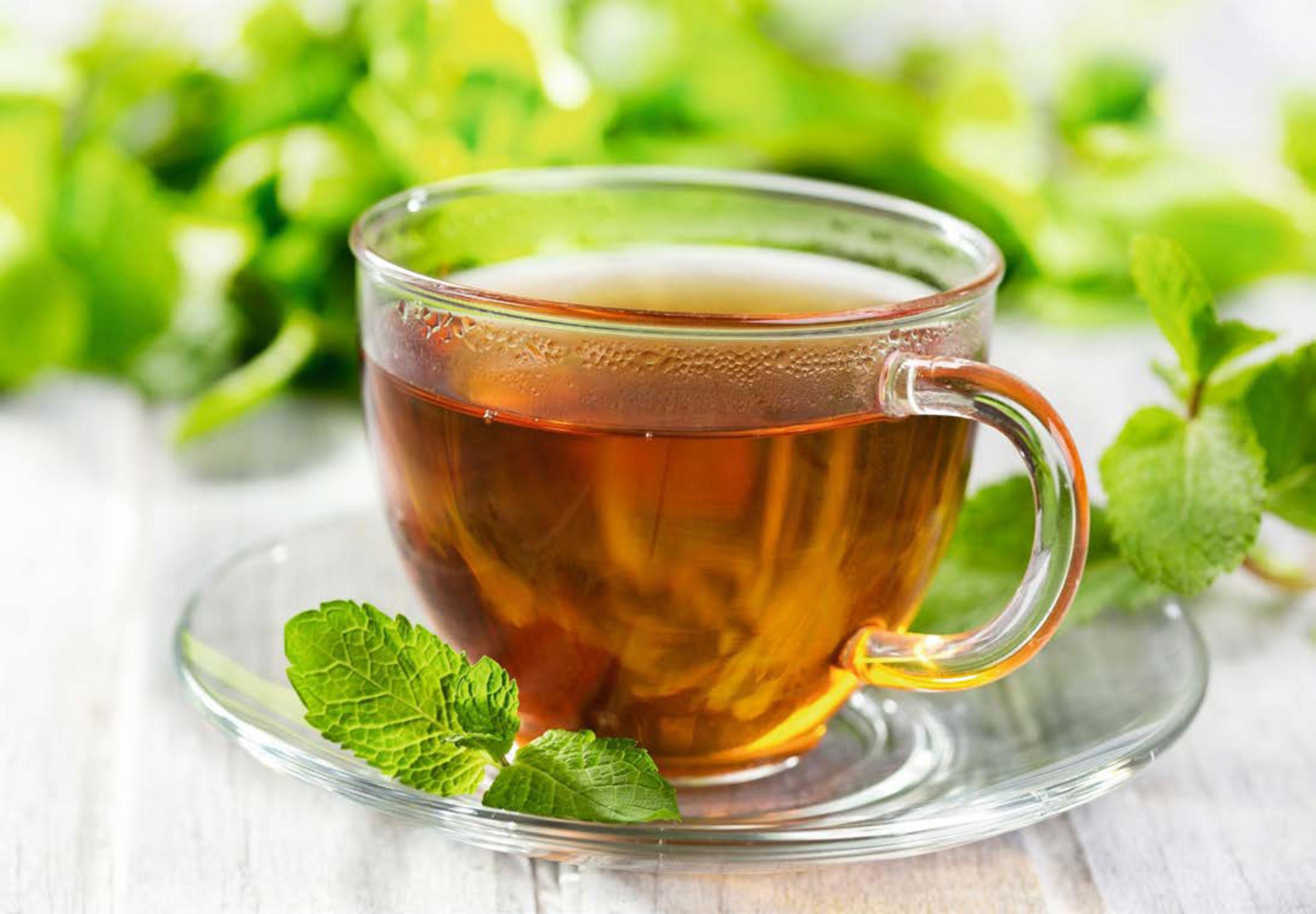 Una taza de té marrón en vidrio transparente, con platillo y hojas de menta. El fondo es una superficie de madera borrosa.