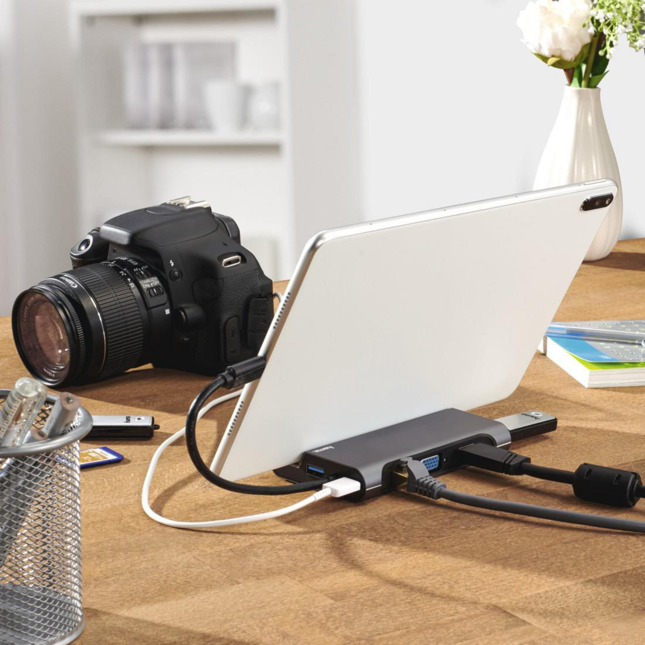 Un tablet collegato a un hub con cavi. Una fotocamera e una penna su una superficie di legno.