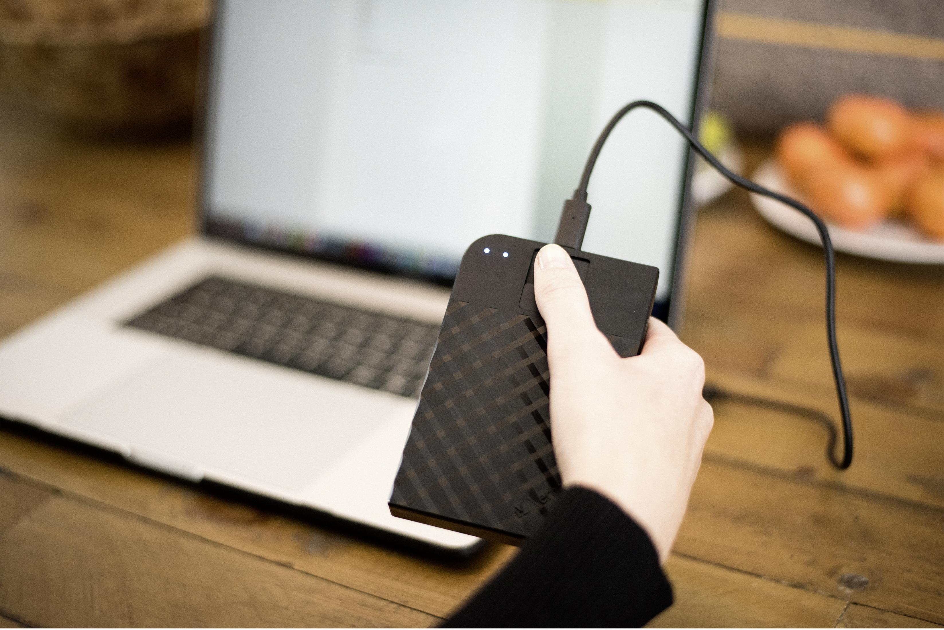 Ręka trzymająca czarny przenośny dysk twardy podłączony do laptopa kablem USB.