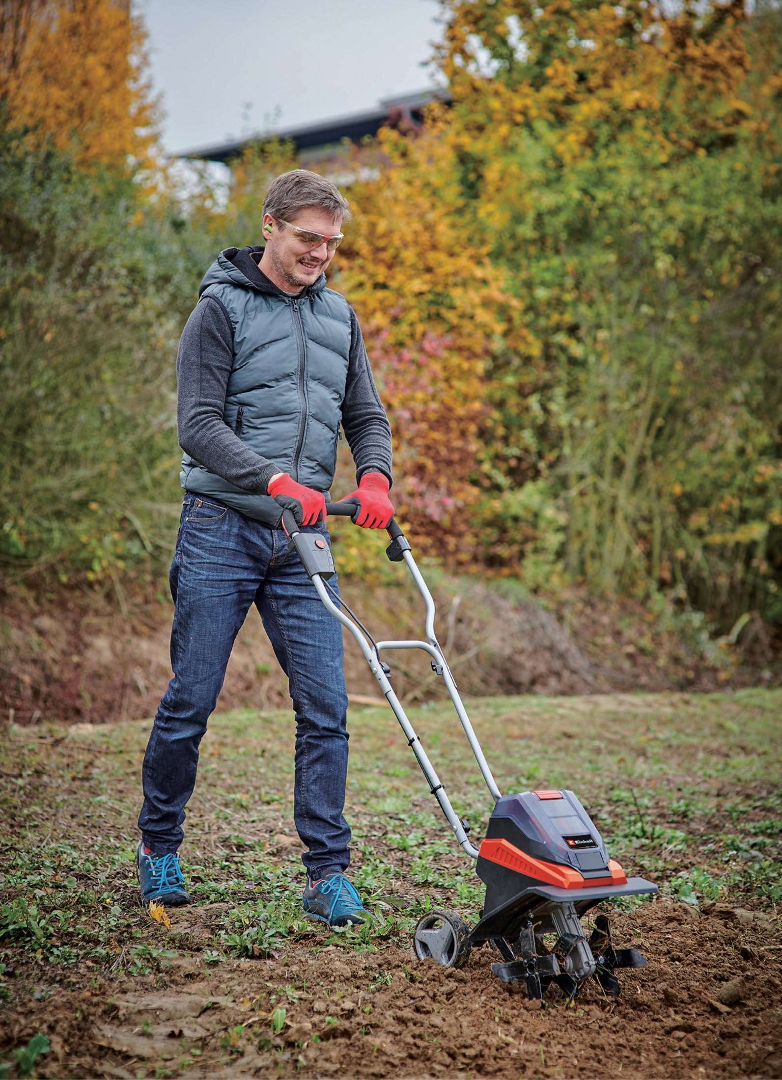 Mann in Jeans und Weste, der einen Bodenfräser auf einem Feld benutzt, umgeben von Bäumen.