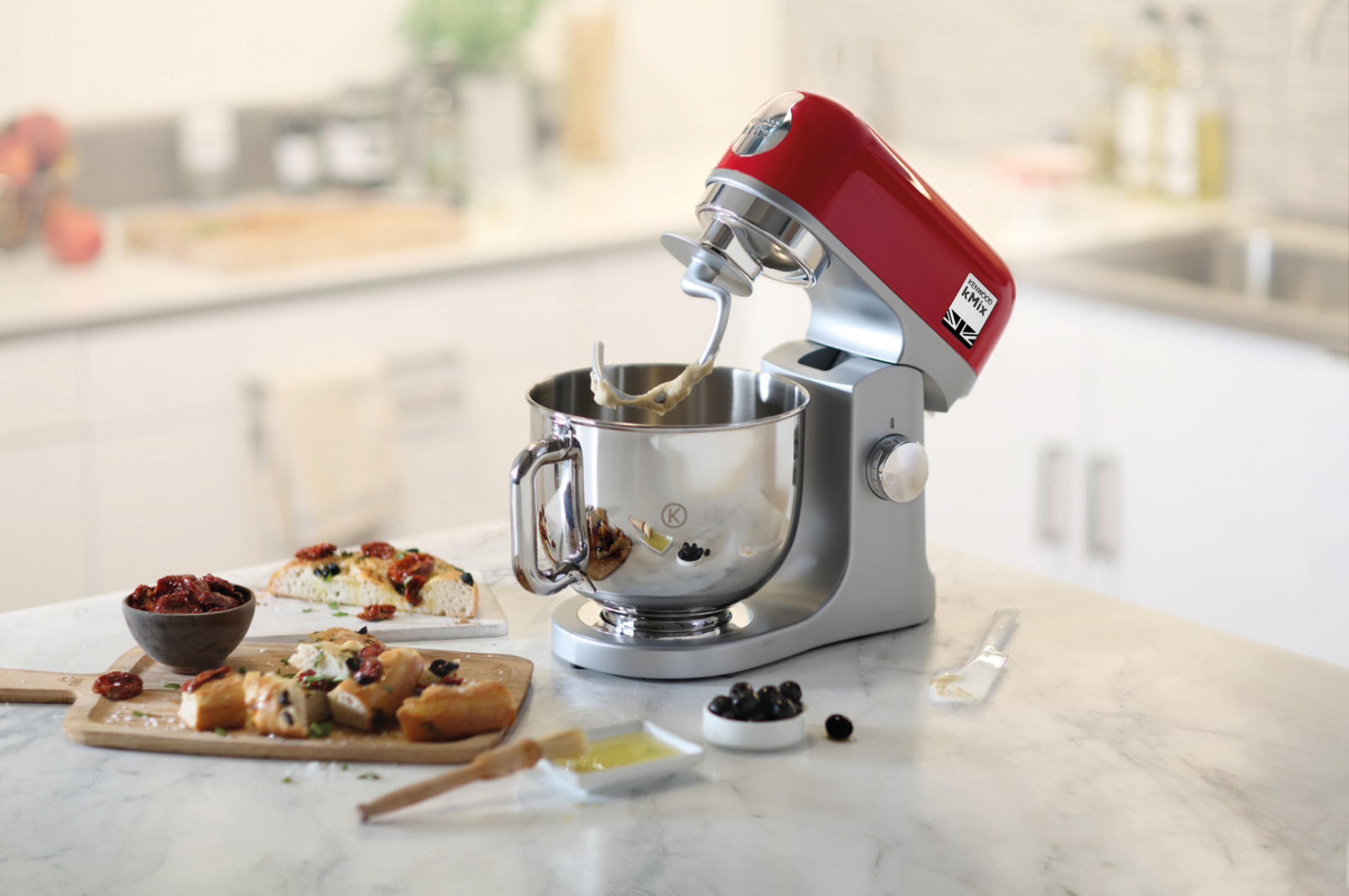 Roter Küchenmixer mit Schüssel. Essen auf dem Tisch: Brot, Oliven, Tomaten und Öl.