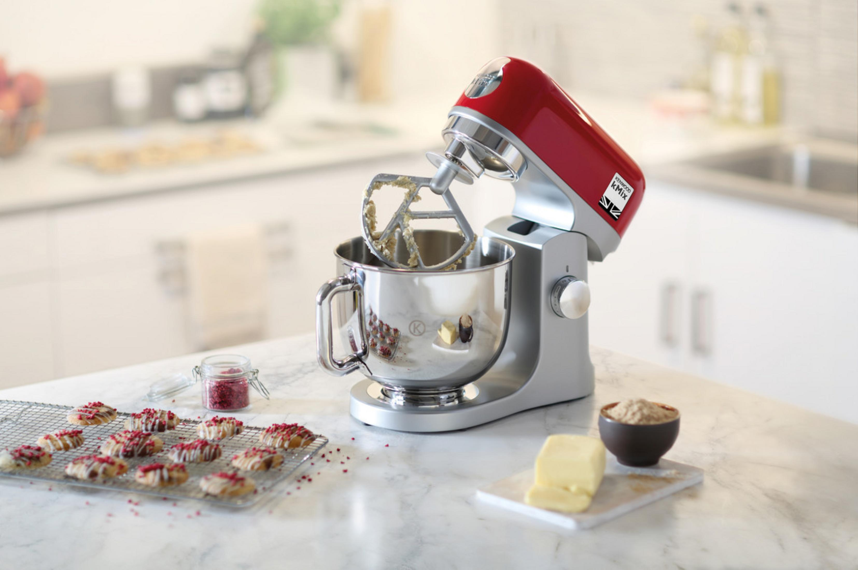 Roter Küchenmixer mit silberner Schüssel, mit Zutaten und gebackenen Keksen auf einer Marmortheke.