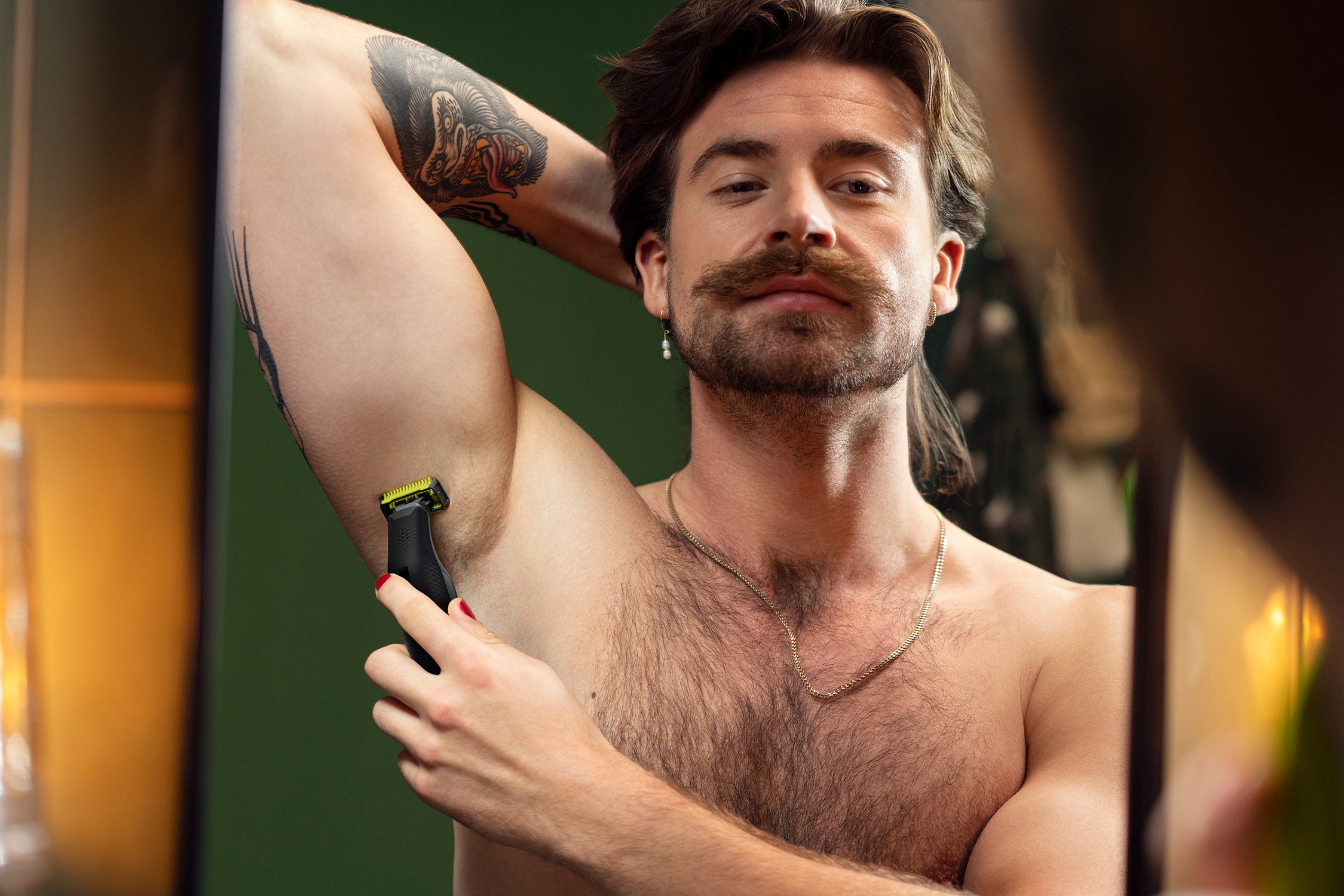Mężczyzna goli pachę trymerem przed lustrem. Ma wąsy i tatuaż.