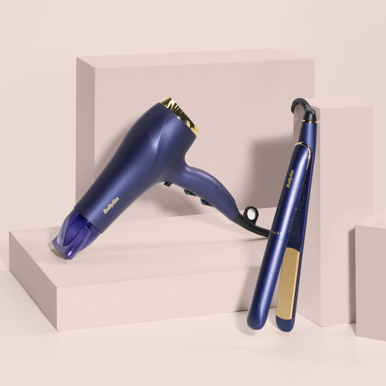 Suszarka do włosów i prostownica, obie granatowo-złote, na białej platformie.