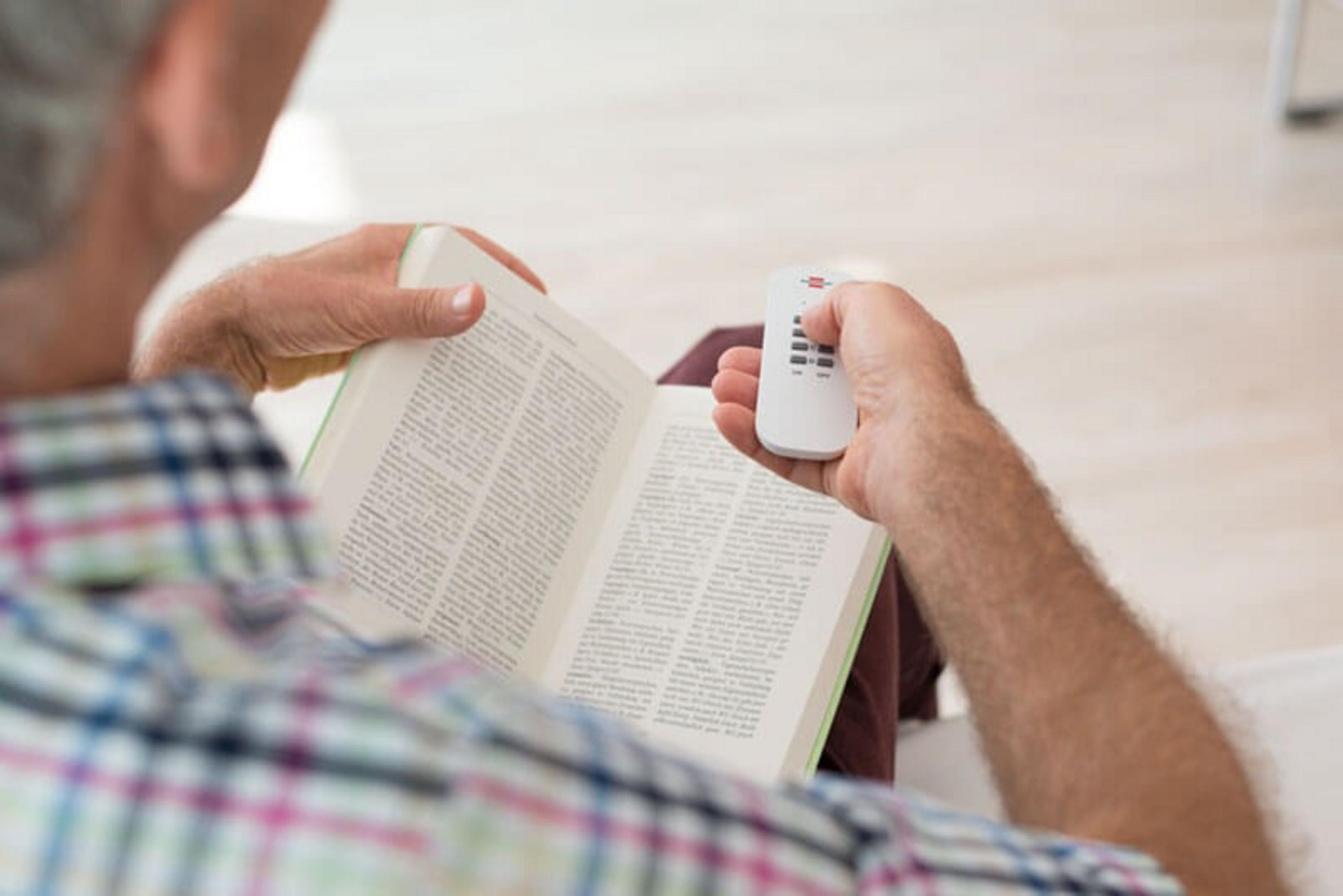 Mann liest ein Buch und hält eine Fernbedienung. Die Ansicht ist von hinten.