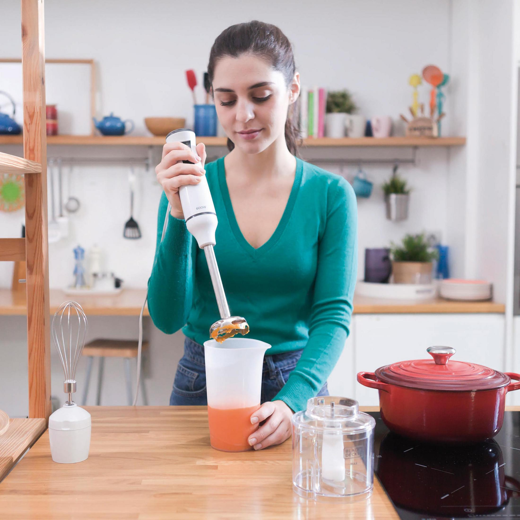 Kobieta używająca blendera ręcznego w przezroczystym naczyniu, z składnikami i akcesoriami kuchennymi.