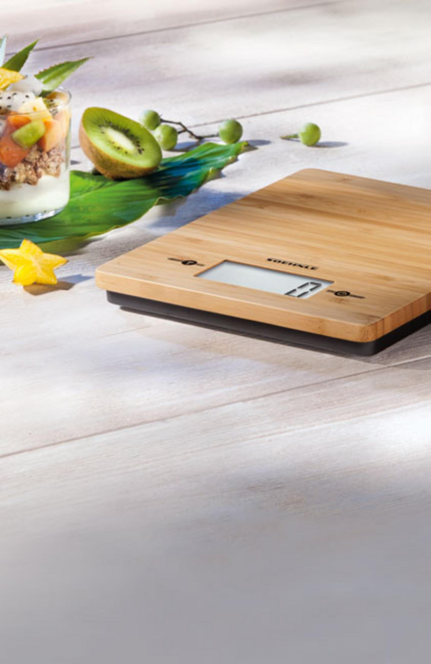 Eine digitale Küchenwaage aus Holz mit Obst und Joghurt im Hintergrund, auf einem Holztisch.