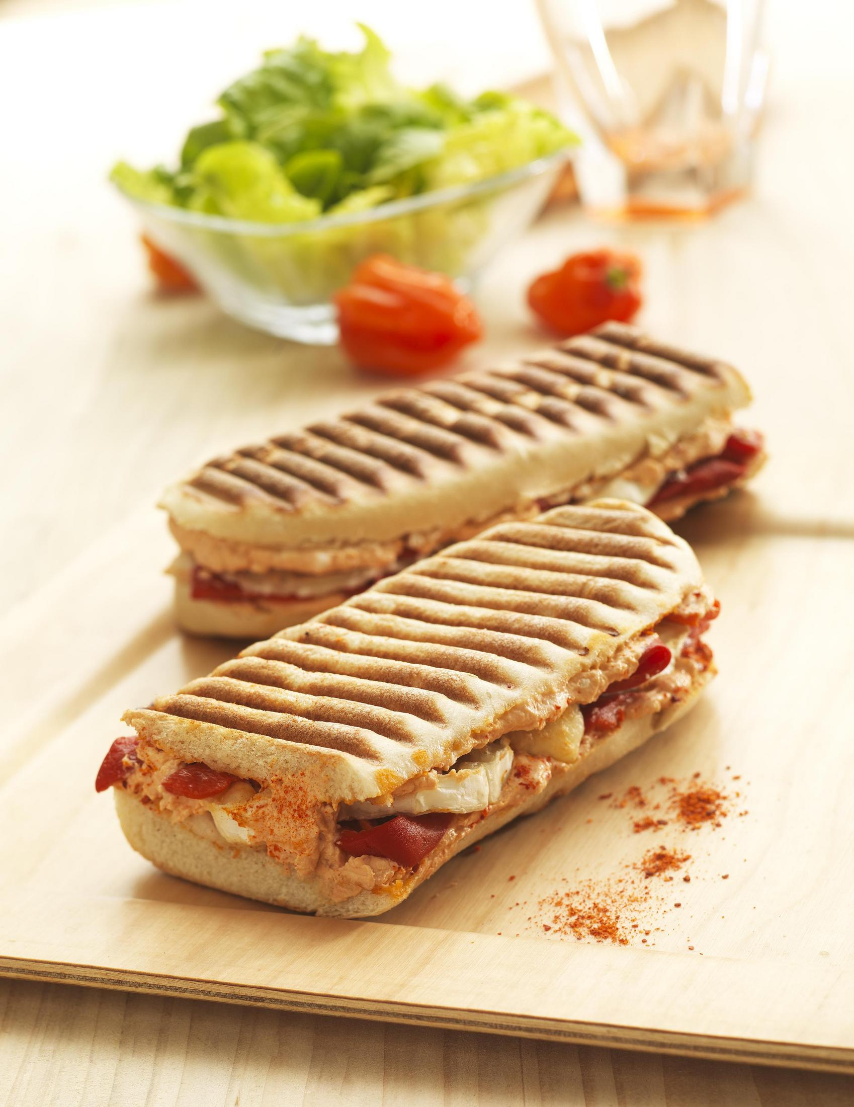 Dwie kanapki panini na drewnianej tacy z sałatą i pomidorami w tle.