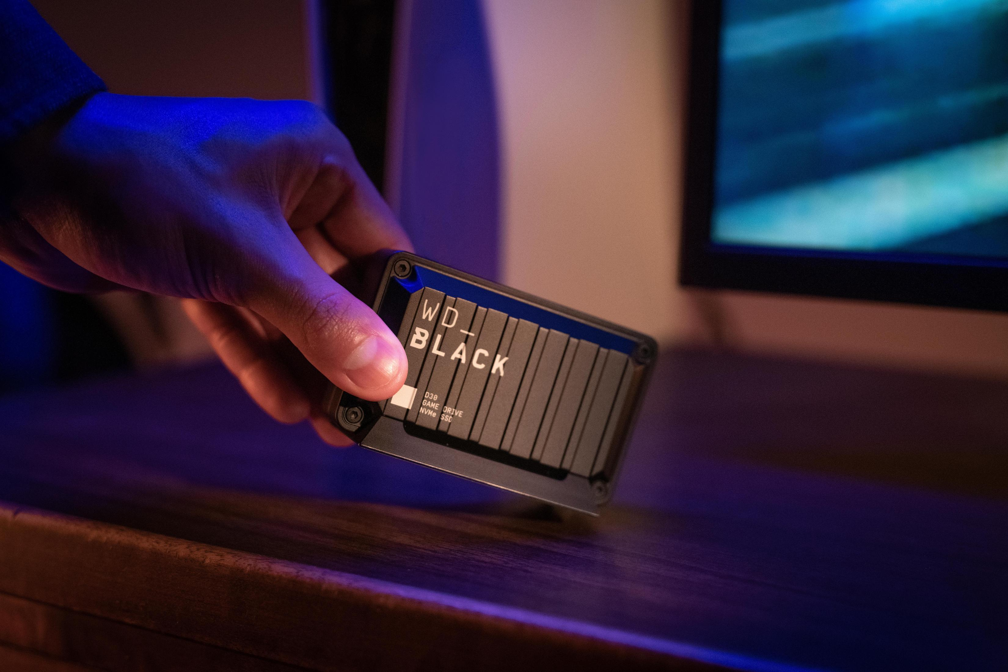 Dłoń trzymająca czarny zewnętrzny dysk twardy WD_BLACK w pobliżu monitora komputera.