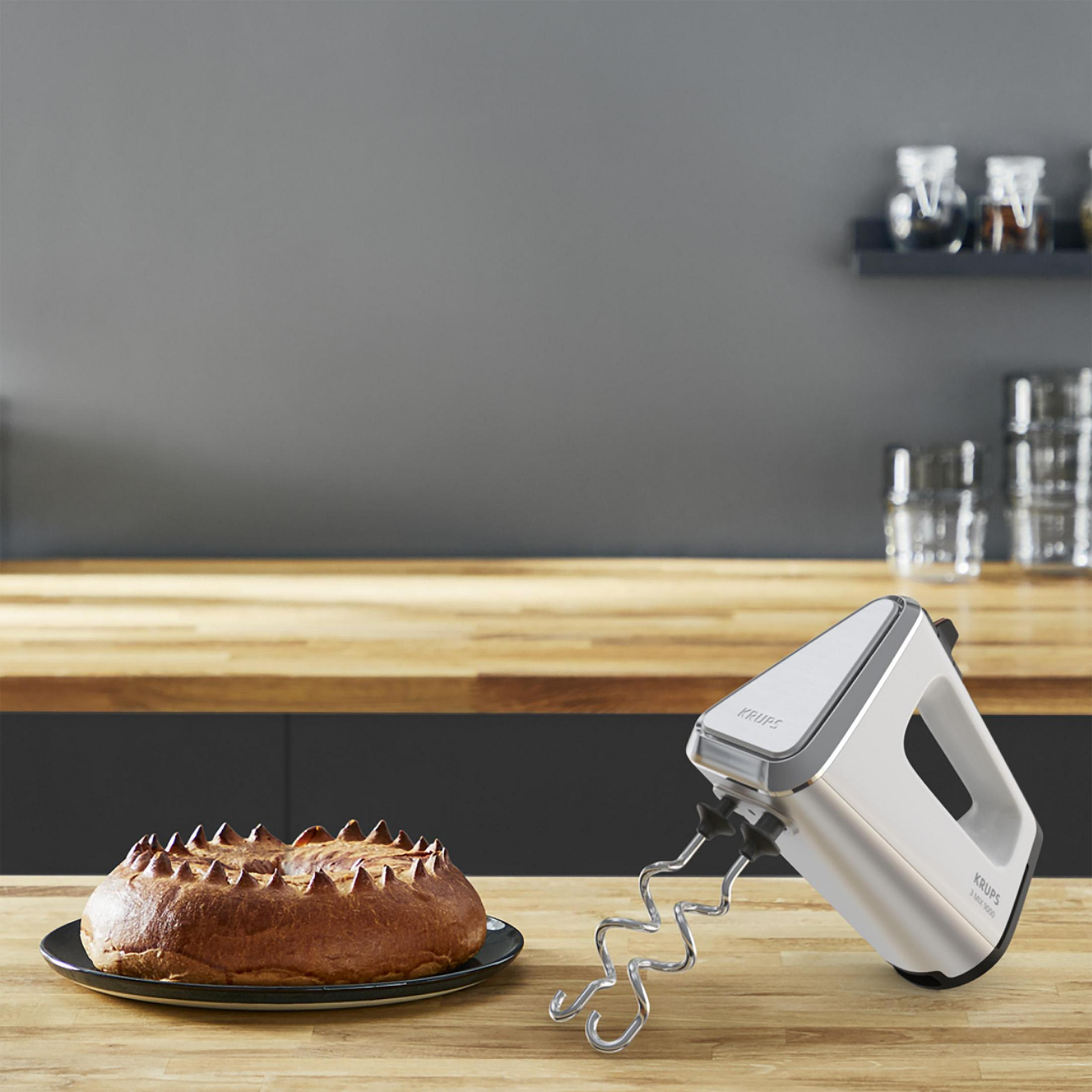 Handmixer neben einem Kuchen. Der Mixer ist silber und weiß, der Kuchen ist dunkel mit Glasur.