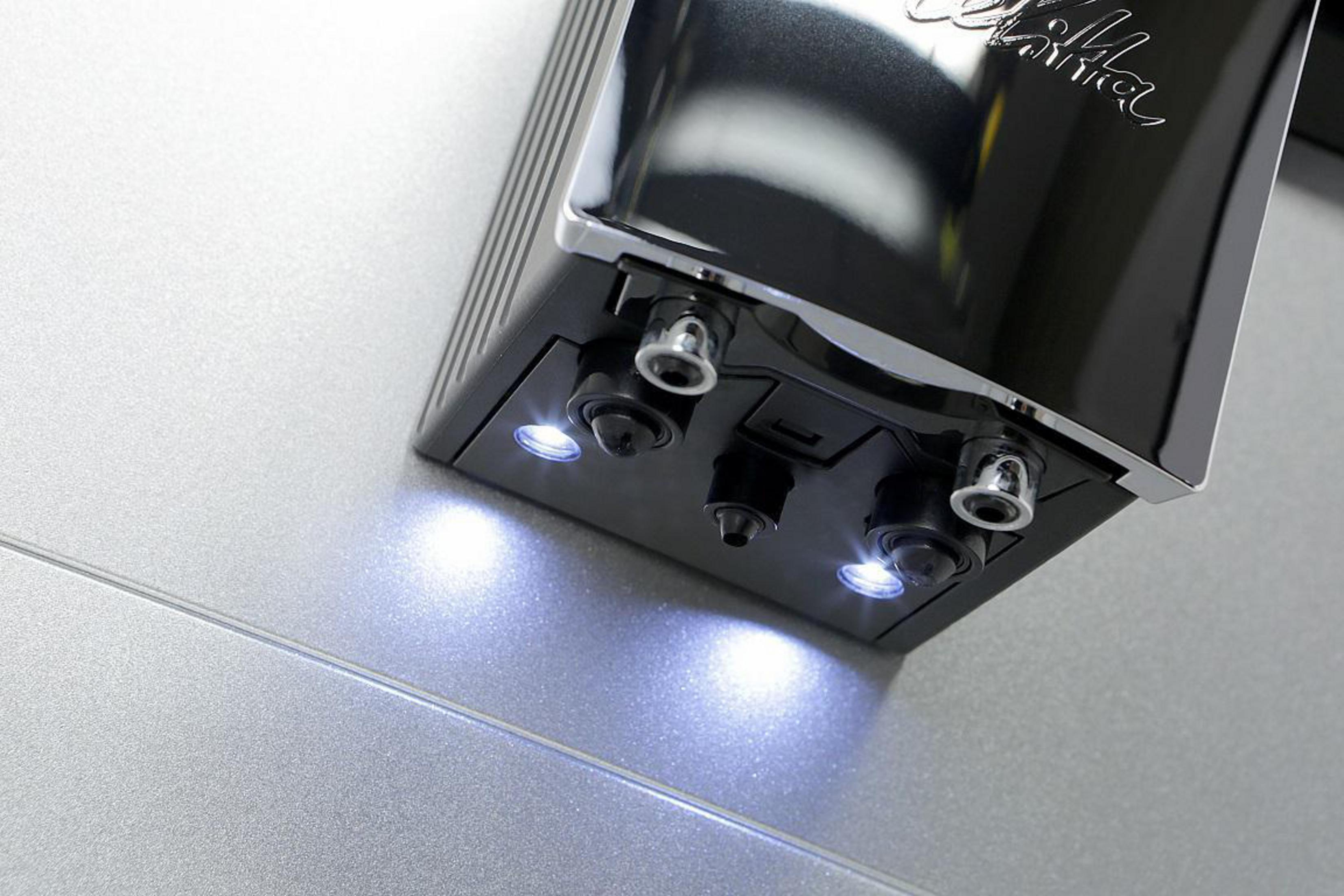 Máquina de café negra y plateada, primer plano. Luces brillan. Parte superior con botones y boquillas.