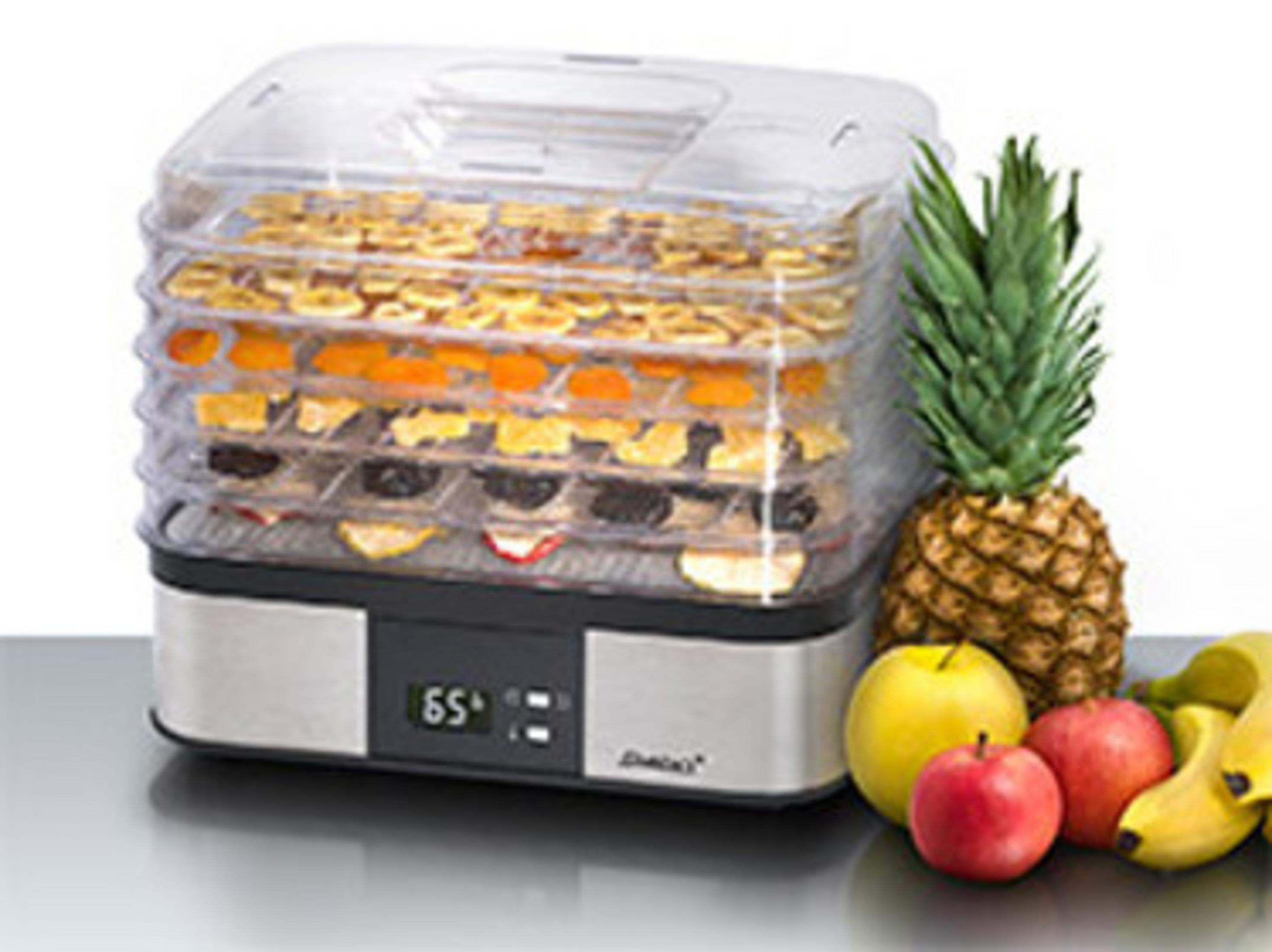 Ein Dörrautomat mit Tabletts voller getrockneter Früchte. Daneben eine Ananas, Äpfel und Bananen.