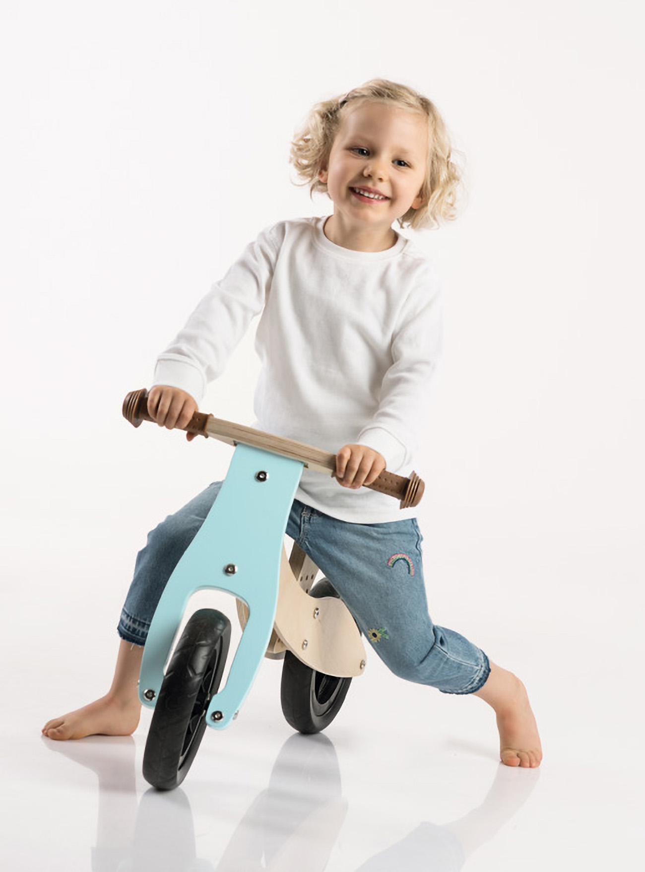 Un bambino sorride e guida una bici senza pedali di legno blu su sfondo bianco.