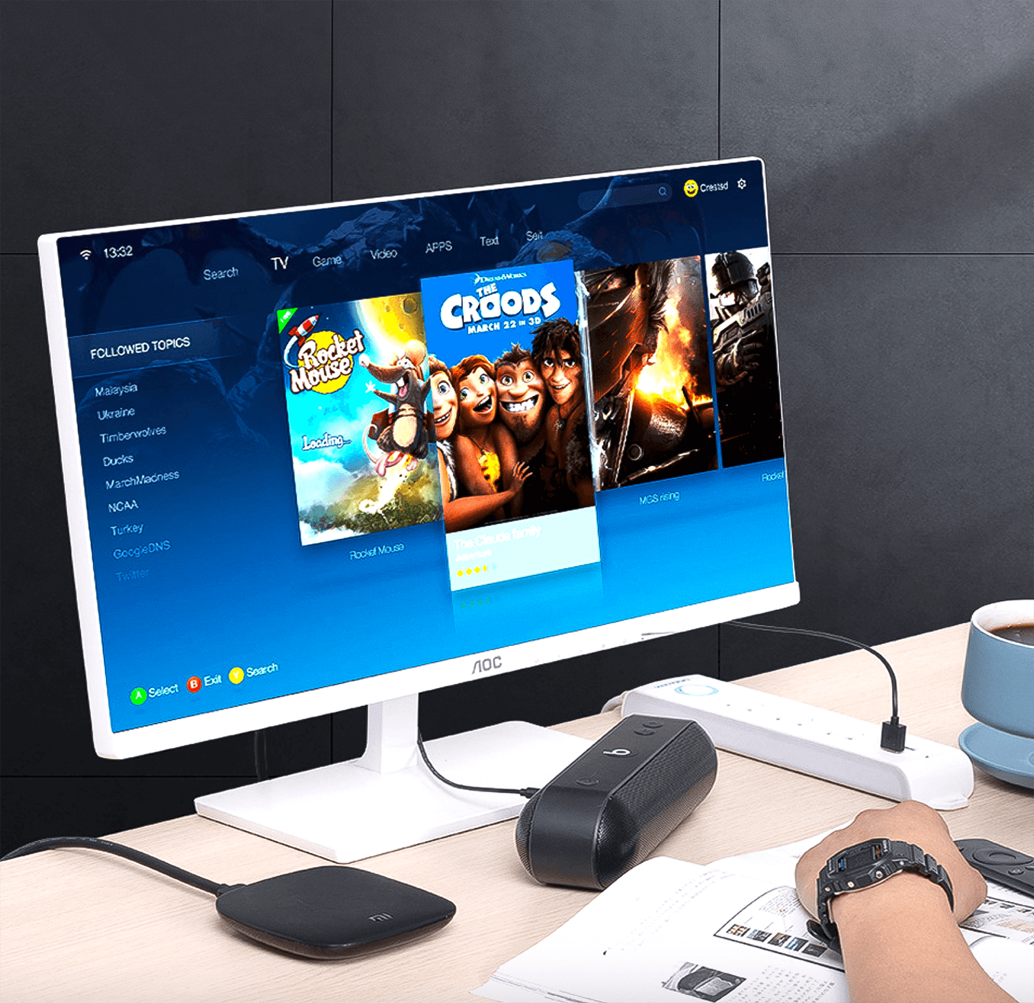 Monitor komputera wyświetla filmy i media społecznościowe. Obok głośnik i dłoń z zegarkiem.