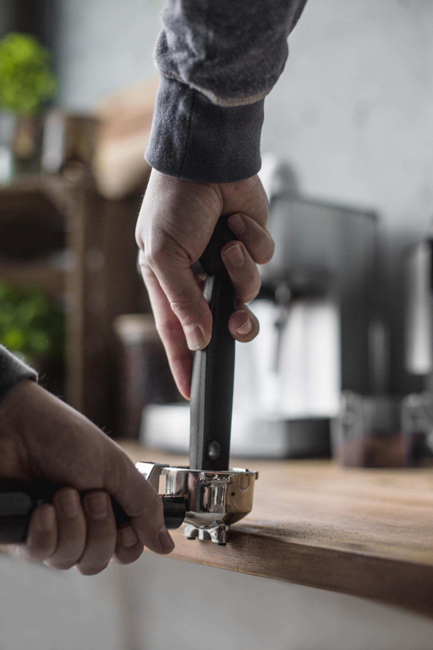 Ręce trzymają czarny portafiltr. Portafiltr jest umieszczany w ekspresie do kawy, na drewnianej powierzchni.