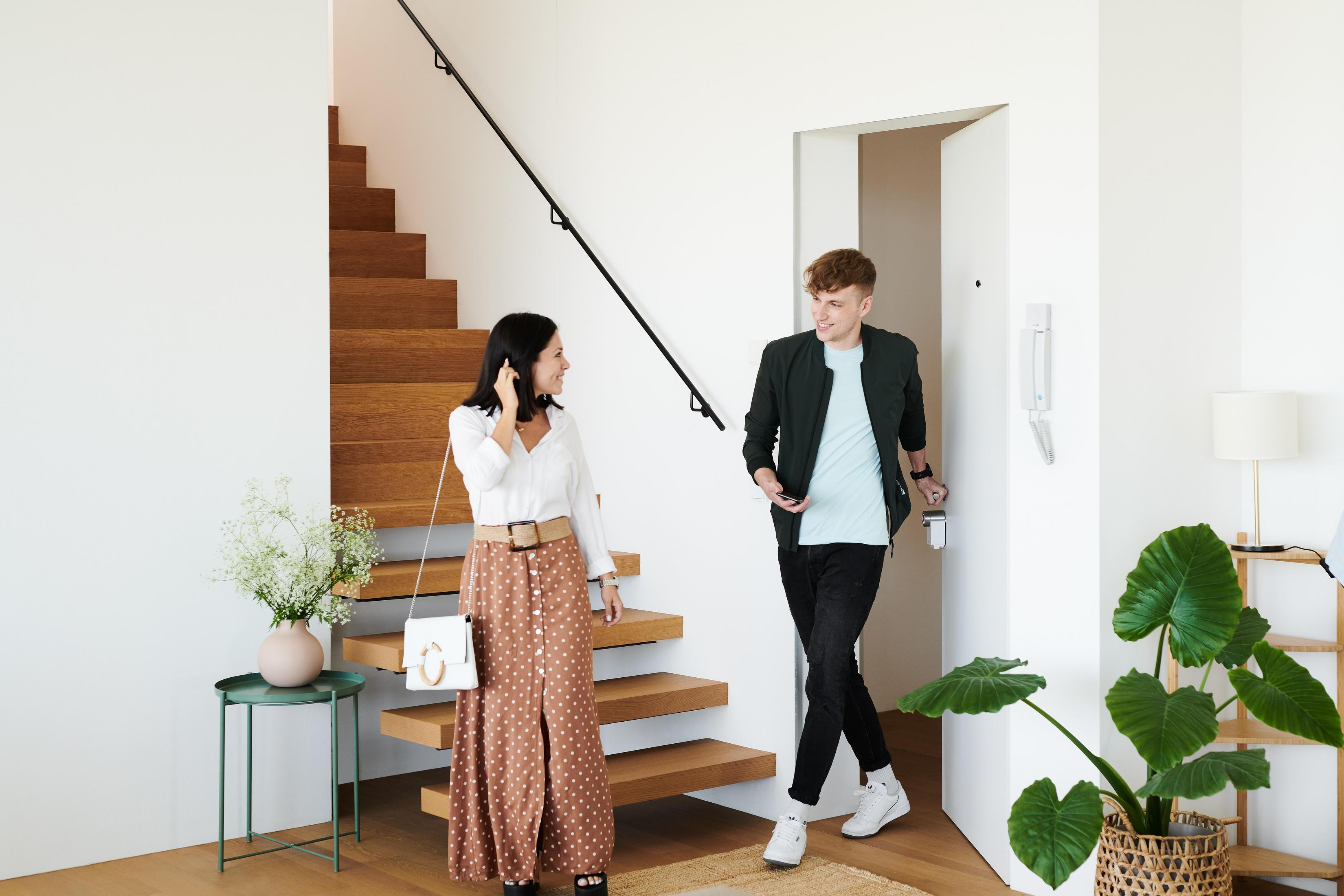 Una pareja entra en una casa. Una escalera y una planta están en el fondo.