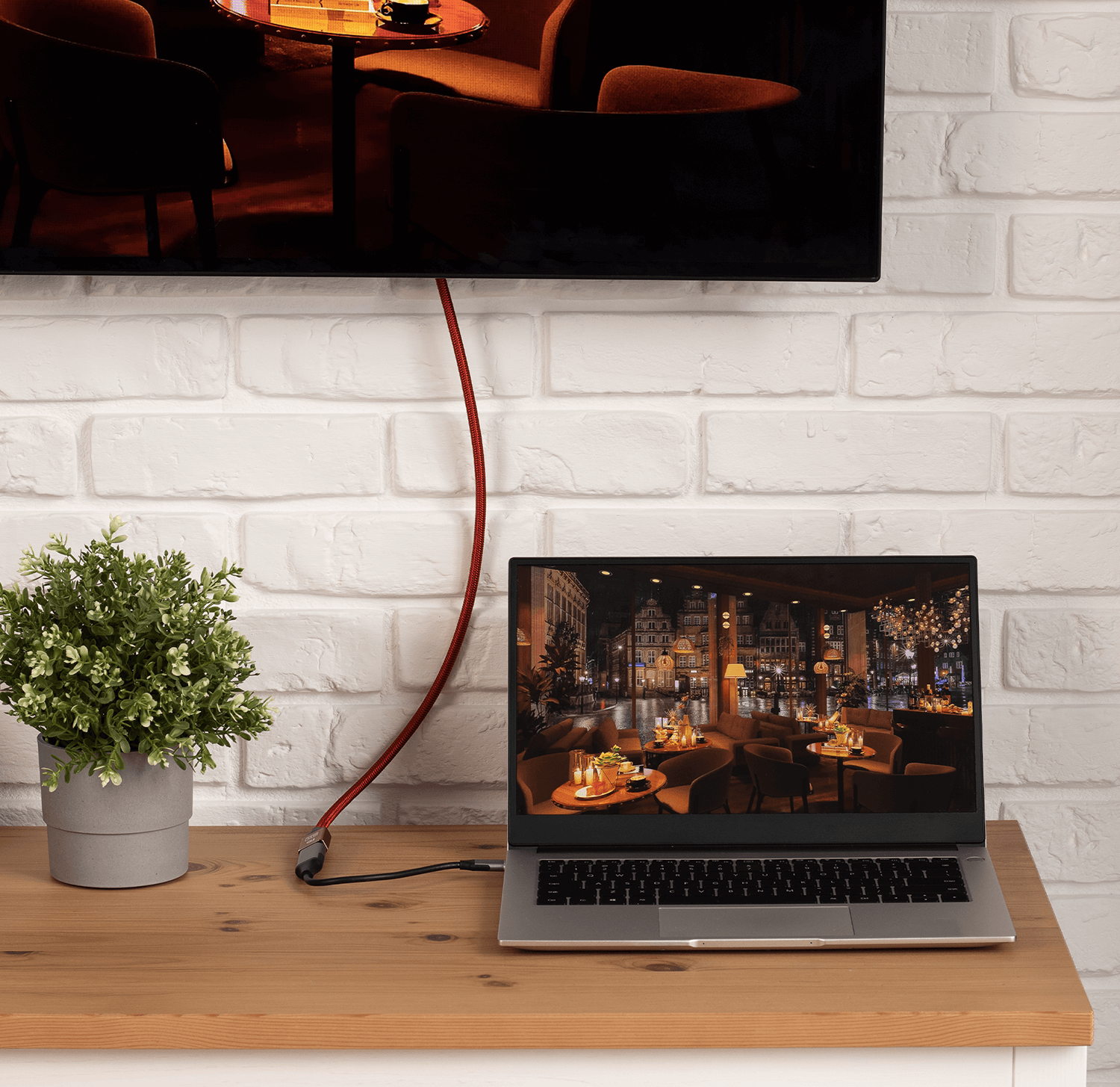 Laptop wyświetlający scenę restauracji podłączony do telewizora, umieszczony na drewnianym stole. Tło z ceglanej ściany.