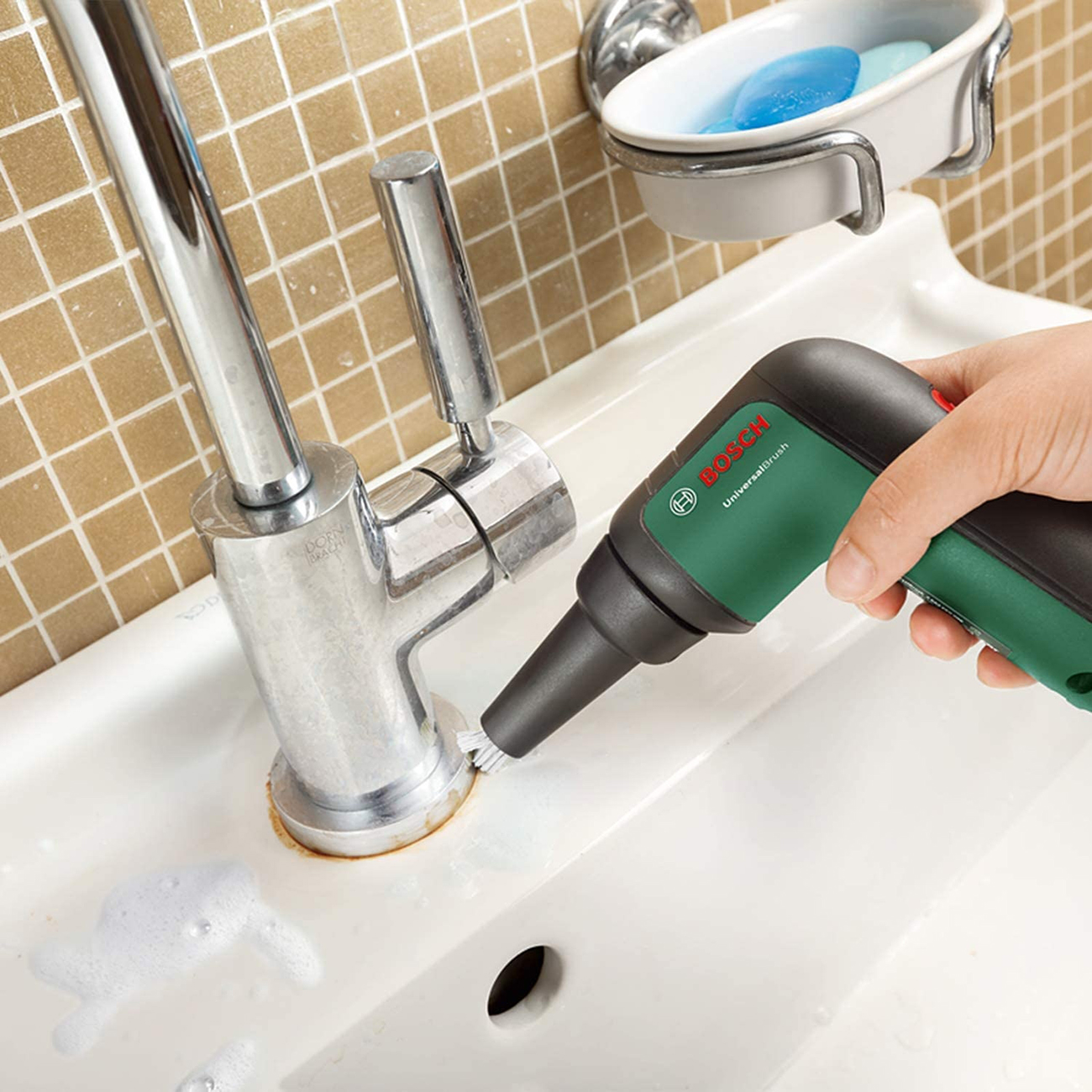 Eine Person reinigt einen Wasserhahn mit einer Bosch UniversalBrush. Seife und Waschbecken sind sichtbar.