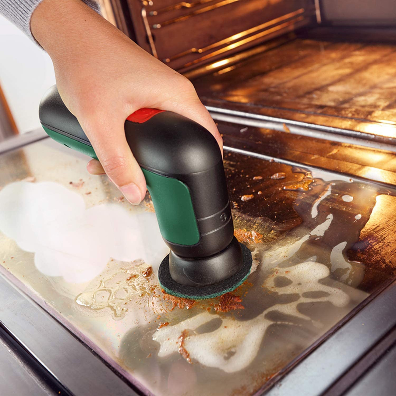 Eine Hand reinigt einen Ofen mit einem elektrischen Scheuergerät. Der Ofen ist verschmutzt. Die Hand ist im Fokus.