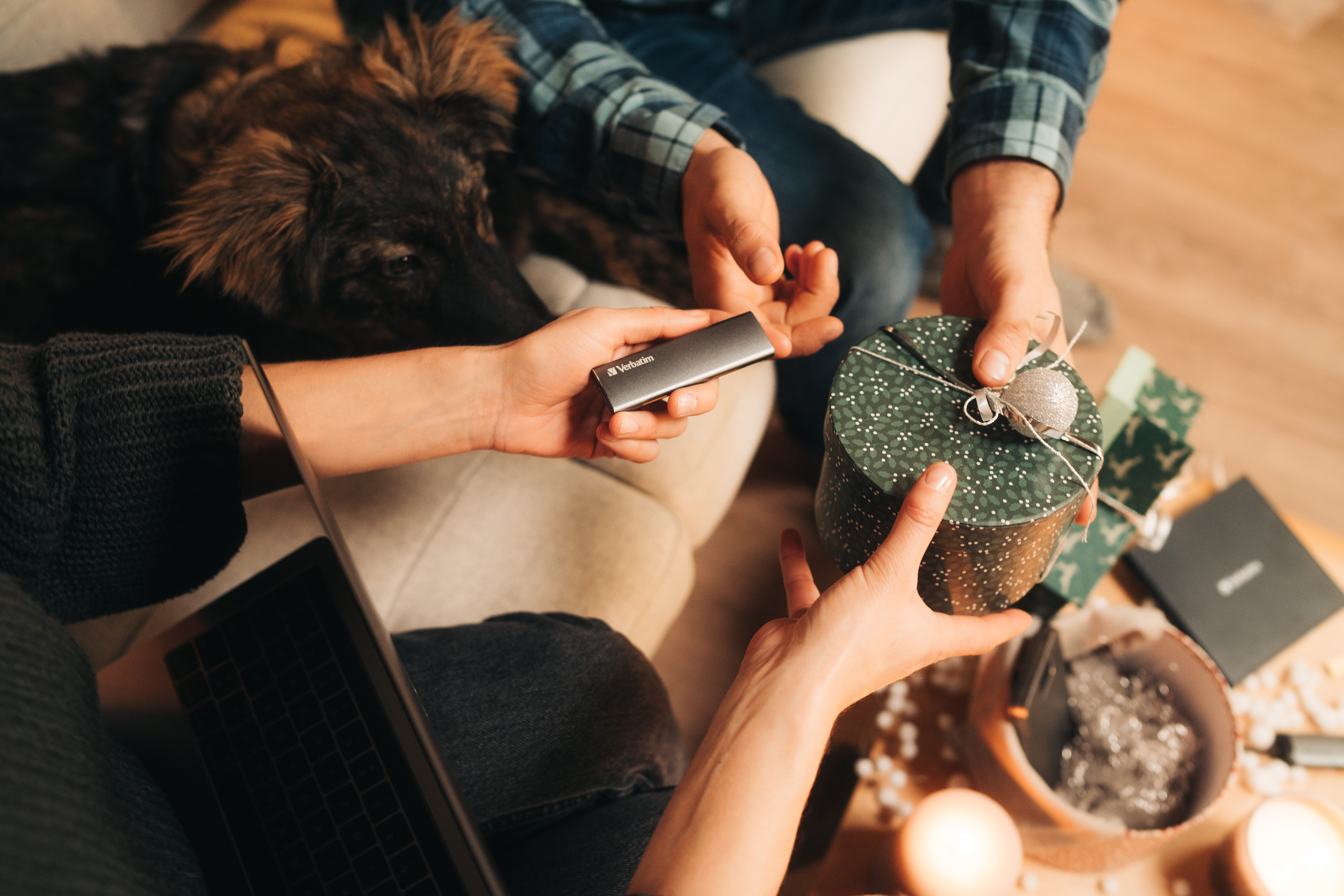 Dwie osoby wymieniające się prezentami. Jedna trzyma dysk Verbatim, druga okrągły zielony prezent. Pies w tle.