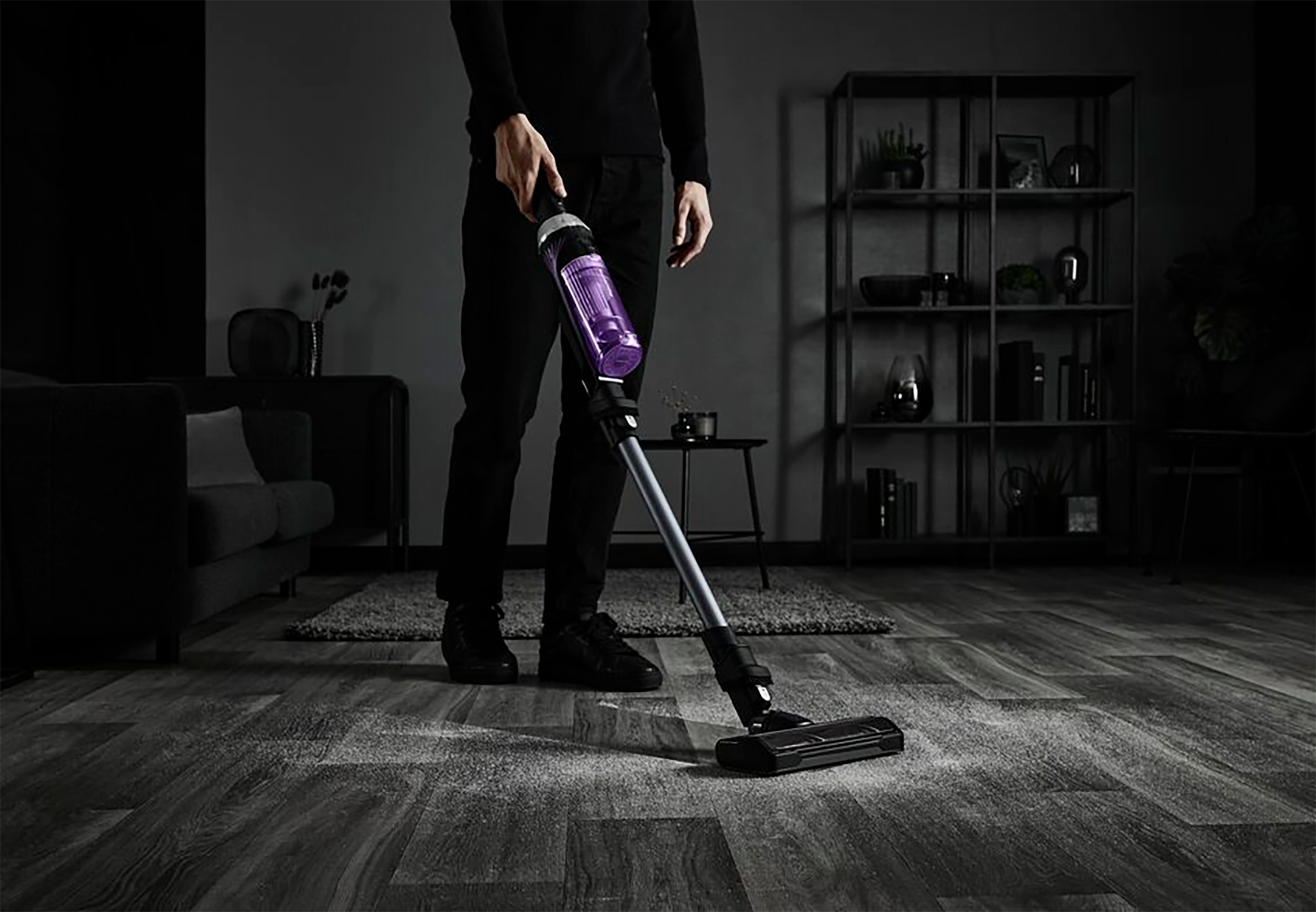 Una persona in nero aspira un tappeto con un aspirapolvere viola. La polvere è visibile. Stanza buia.