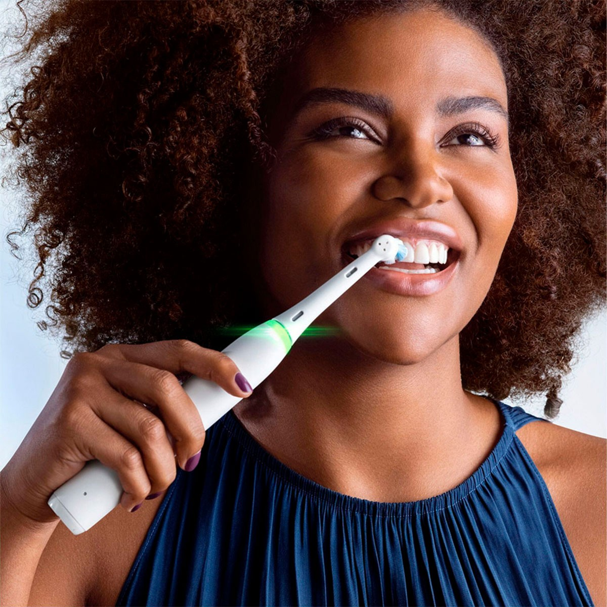 Mujer cepillándose los dientes con un cepillo eléctrico. Sonriendo, top azul.